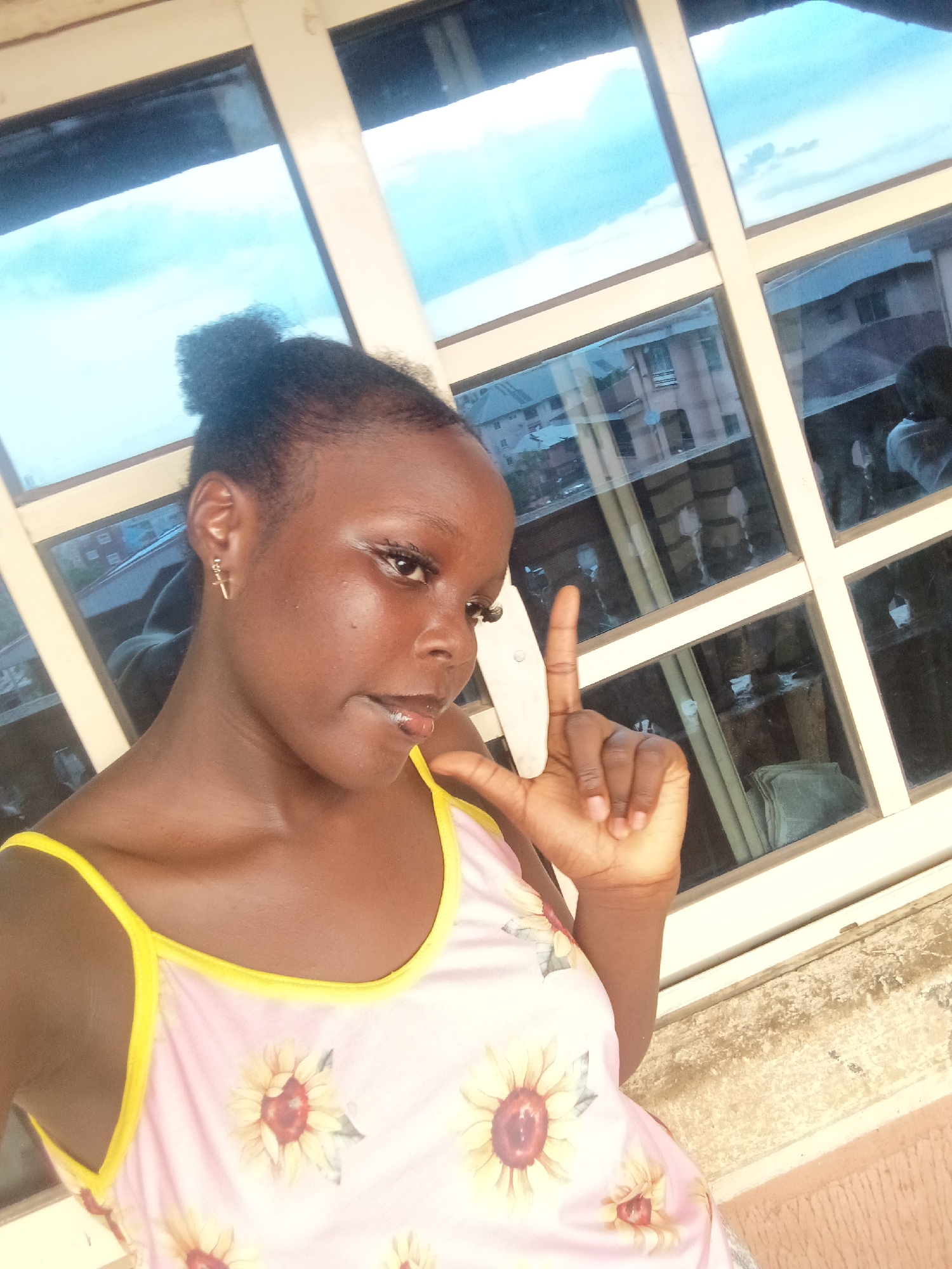 The image displays a young Black female, appearing to be in her late teens or early twenties, positioned indoors against a window in the city of Onitsha, Nigeria. Her dark hair is gathered in a high bun. She is wearing a light pink sleeveless top with yellow trim and sunflower patterns. A small gold cross-shaped earring is visible in her left ear. Her lips have a glossy finish. Her right hand is raised, making a gesture with her index finger and thumb extended, partially obscuring a white object behind it.
The background consists of a multi-paned window with cream-colored or light beige frames. The window panes are reflective and exhibit a blue tint. Through the upper panes, a bright, cloudy sky is discernible. Reflections in the lower and middle window panes show parts of multi-story buildings, featuring light-colored facades and balconies, typical of urban residential or commercial architecture in a Nigerian city. A textured, light brown surface, likely a wall or window sill, is visible below the window frame.