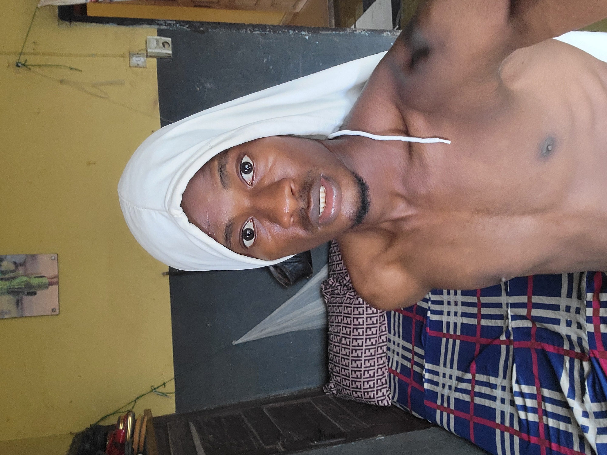 A young Black man with a white cloth covering his head is positioned in the foreground of the image, looking directly at the camera. He is shirtless and appears to be in a bedroom. Behind him, a bed is visible with a patterned comforter and pillow. The room's walls are painted yellow and dark gray, with electrical outlets and a picture hanging on the yellow wall. The overall setting suggests a domestic environment in the city of Onitsha, Nigeria.