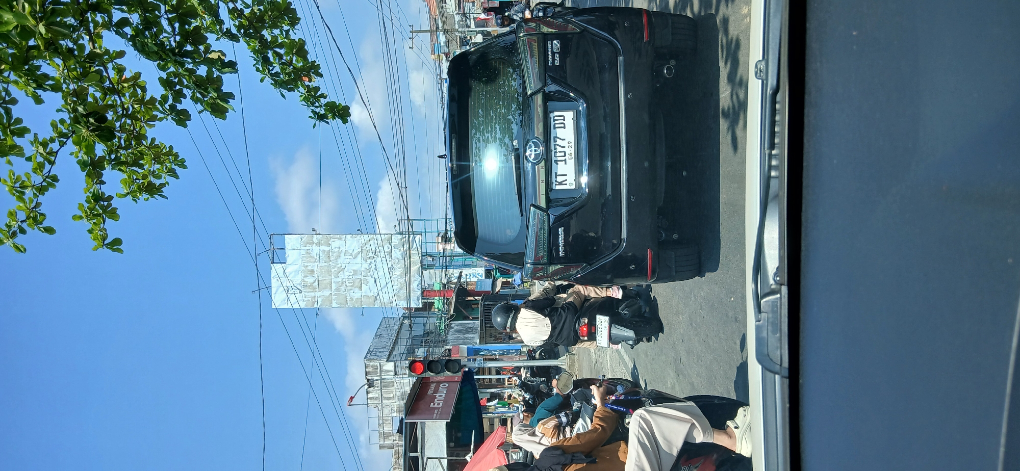 This is a street-level, eye-level photograph taken on a bright, sunny day in Samarinda, Indonesia, likely at an intersection controlled by traffic lights. The foreground is dominated by the interior of a vehicle, specifically the windshield and dashboard. The main subject is a black Toyota Innova SUV from its rear, stopped in traffic. The license plate reads "KT 1077 DD" with an expiry date of "06.29".
To the left of the SUV, two individuals on a motorcycle are visible. The rider is wearing a helmet and a dark jacket with a light-colored back panel. The passenger, a woman wearing a hijab and a brown jacket, is partially visible. Another motorcycle is also stopped in traffic, with its rider wearing a helmet.
In the background, there are buildings with faded billboards. One prominent billboard is tall and metallic, appearing weathered. Another building to the left has a red sign that reads "PERTAMINA Enduro". A traffic light is visible, currently showing a red signal. Overhead, power lines crisscross the sky, and a green tree with large leaves hangs into the frame from the top right. The overall scene depicts a typical urban traffic situation in Indonesia.