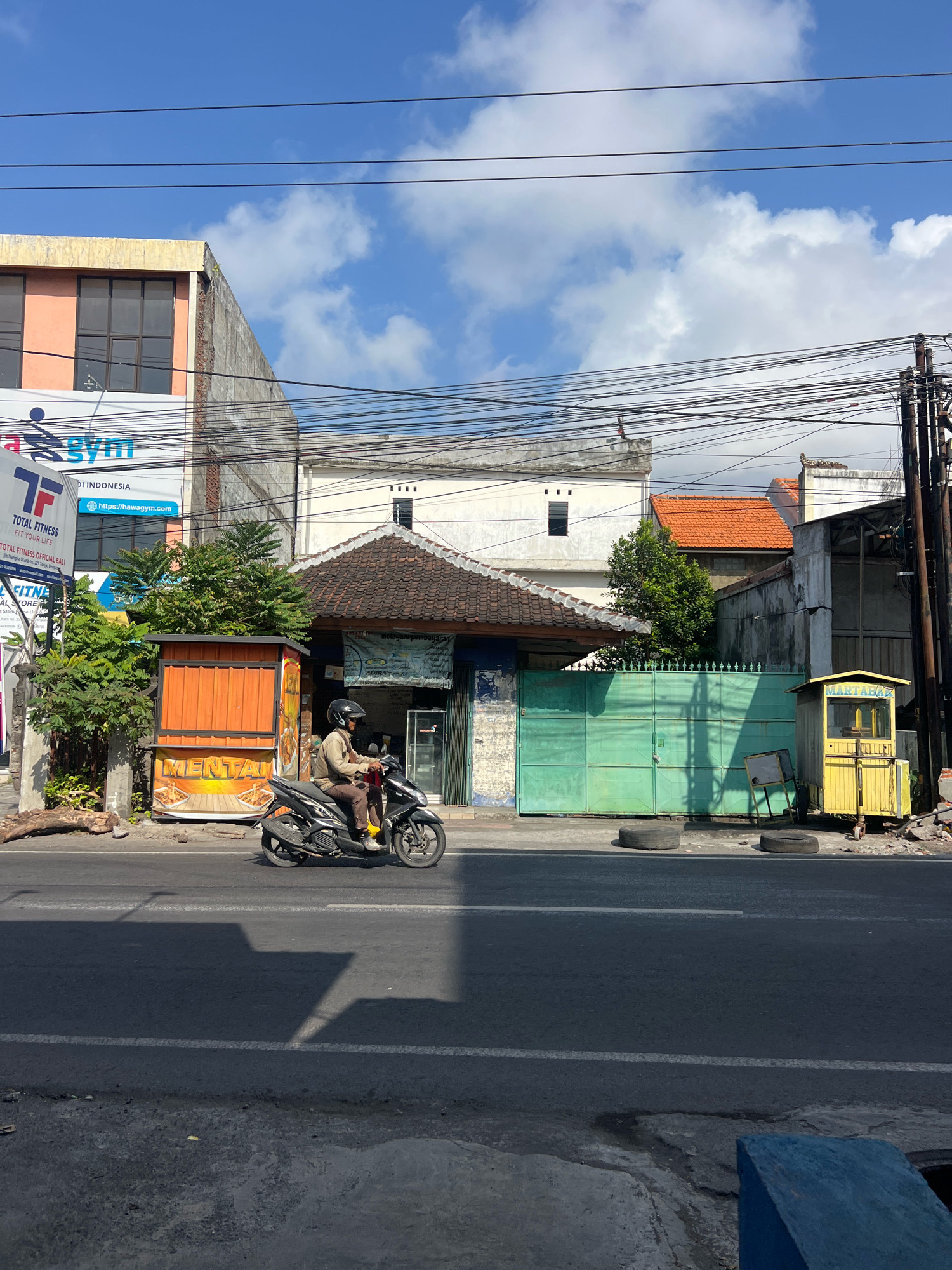The image depicts a street scene in Denpasar, Indonesia, on a sunny day with scattered clouds. In the foreground, a dark asphalt road runs horizontally across the frame. A person wearing a helmet and a light-colored jacket is riding a black motorcycle in the right lane, moving from left to right. The motorcycle has its headlights on. On the left side of the road, there is a narrow orange food stall with the word "MENTAI" prominently displayed. Behind the stall, lush green trees grow. Further back, several buildings are visible. To the left, a multi-story building with a pink facade and dark windows stands. To its right, a white building with a tiled roof is partially obscured by wires. In the middle ground, a blue-green metal gate blocks access to a property. To the right of the gate, a small yellow booth with the word "MARTABAK" is situated, next to a pile of debris. Utility poles and a dense network of overhead wires dominate the upper portion of the image, indicating an urban environment. The overall scene suggests a typical day in a bustling Indonesian city, with commerce and daily life unfolding along the roadside.