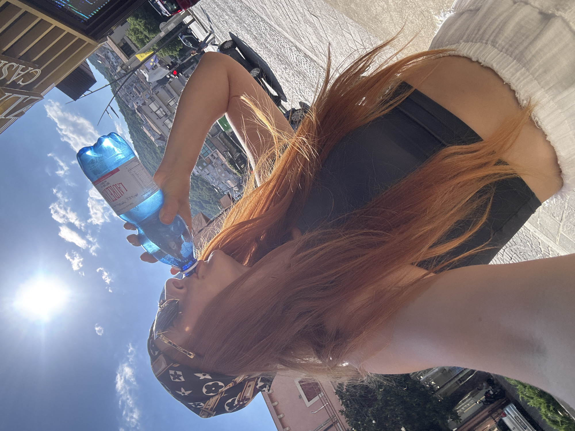 A person with long, red hair and wearing a black crop top, white pants, sunglasses, and a patterned bandana is drinking from a blue plastic bottle of water. The person is standing outdoors on a sunny day, with the sun shining brightly in the sky. In the background, there are buildings, a street with cars, and a hill with more buildings. The scene appears to be a city street, possibly in Budva, Montenegro, given the architectural style of the buildings and the hilly terrain.