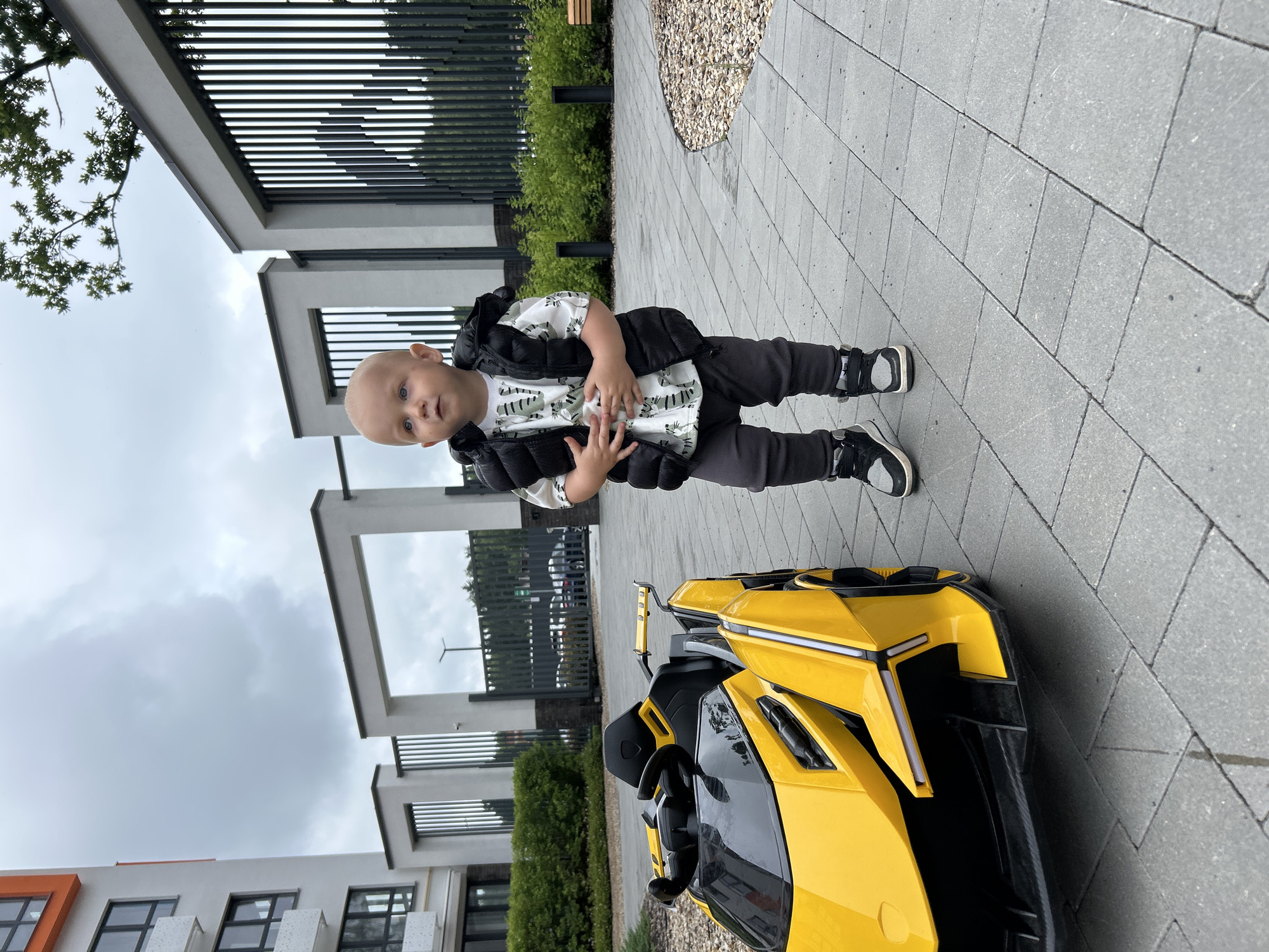 A bald toddler stands on a paved courtyard next to a yellow toy sports car. The toddler is wearing a white t-shirt with a green pattern, a black vest, grey pants, and black and white sneakers. The car is a miniature replica of a Lamborghini. In the background, there is a modern apartment building with white and orange accents, a black metal fence, green bushes, and trees. The sky is overcast. The location is an outdoor area in the city of Kaluga, Russia.