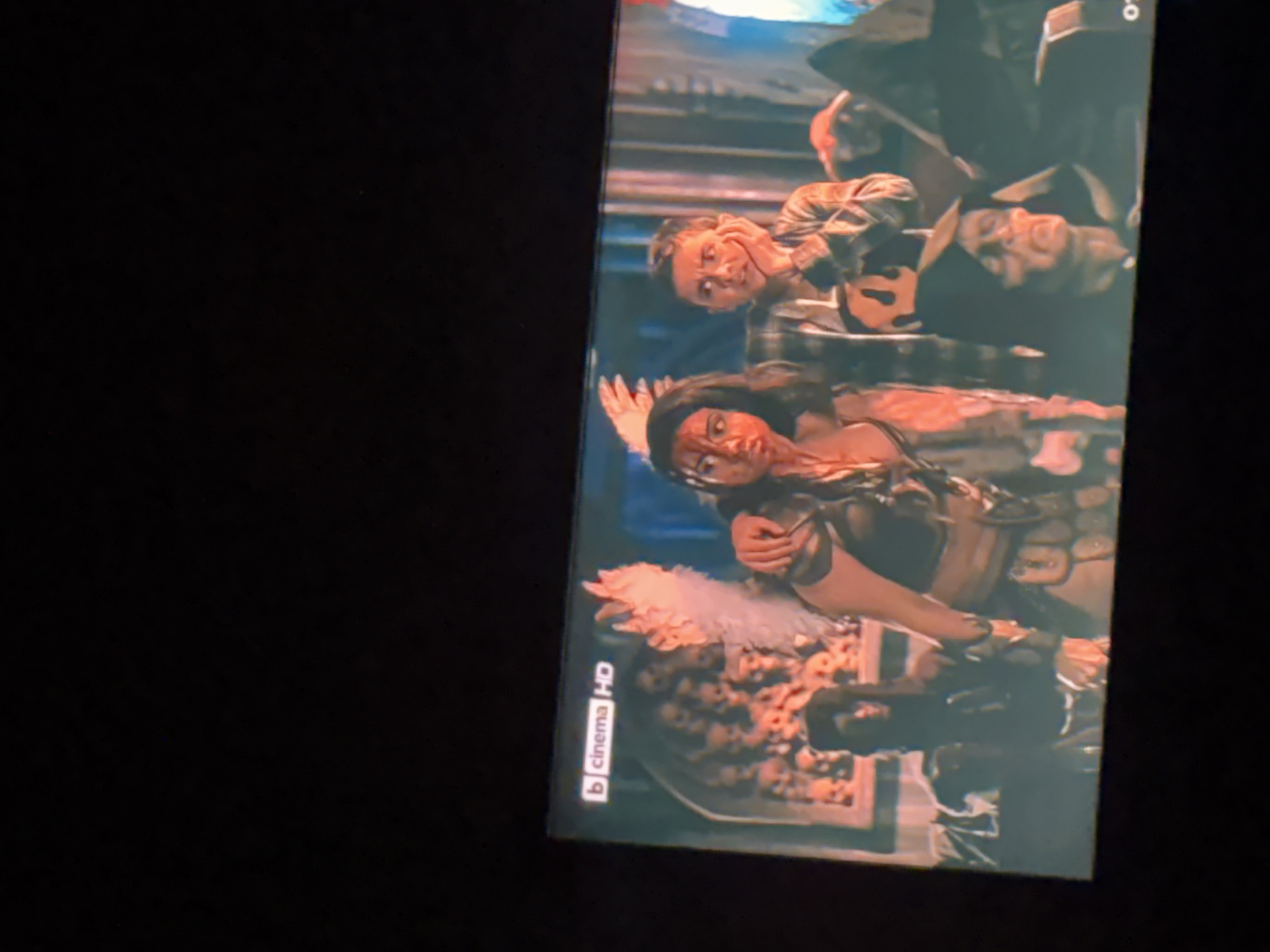 A movie scene is depicted on a television screen. The image shows a group of people in what appears to be a fantasy or post-apocalyptic setting. In the foreground, a woman with dark hair and blood on her face is positioned to the left, wearing an elaborate costume that includes what looks like wings. To her right, a young person in a plaid shirt is looking towards the left with their hand on their chin. Behind them, other figures are visible, some wearing hoods. The background features a wall adorned with numerous skulls. The top left corner of the screen displays the "b cinema HD" logo. The scene appears to be a staged production for entertainment purposes and is not located in Iskar, Bulgaria.
