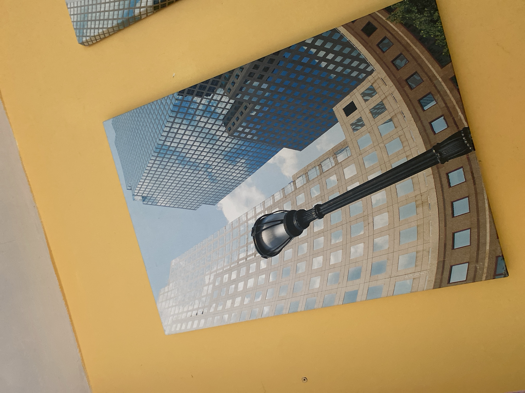 The image shows a framed print of a city scene, featuring modern skyscrapers and a street lamp. The print is displayed on a yellow wall. The skyscrapers are tall, with reflective glass facades. One building in the foreground is a lighter color, with a grid pattern of windows. Behind it, a darker blue skyscraper reflects the sky. A black wrought-iron street lamp is positioned in the lower center of the print, extending upwards in front of the buildings. The sky in the print is a light blue with some clouds. Another framed print with a similar architectural pattern is partially visible to the right of the main print. The wall color is a vibrant yellow, and the ceiling above is a pale gray. The lighting suggests it is daytime. This image does not depict any specific location in Wrocław, Poland.