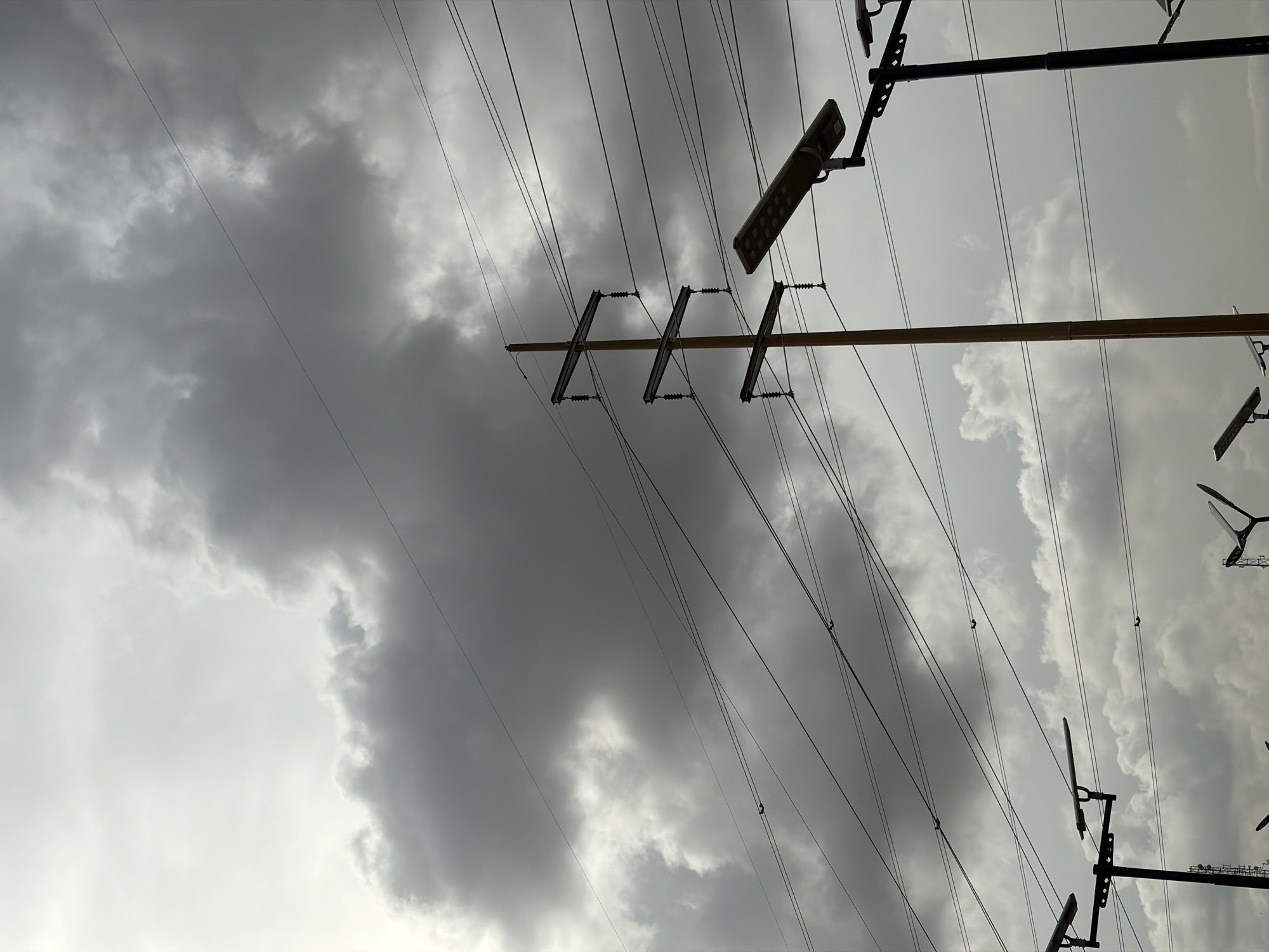 The image presents a complex overhead view of utility infrastructure against a heavily overcast sky. Numerous dark power lines crisscross the frame, extending in various directions and creating a dense, geometric pattern. A prominent wooden utility pole is visible, angled diagonally from the lower-left to the middle-right. It features multiple cross arms equipped with insulators that support many of the power lines.
To the right of the wooden pole, a darker, possibly metal, pole supports what appears to be a modern solar-powered street light. This rectangular fixture, fitted with multiple LED-like elements, is angled downwards. Other similar lighting fixtures or structures are partially visible along the far right edge of the image.
The sky is dominated by thick, dark grey clouds, which are particularly dense and heavy in the upper left and central areas. Lighter patches of grey and white clouds are also interspersed throughout, indicating variations in cloud cover. The overall impression is of an impending storm or a day with very heavy cloud cover. The lighting is dim and diffused, consistent with overcast weather. No people or specific activities are visible in the static scene. There is no discernible text within the image.