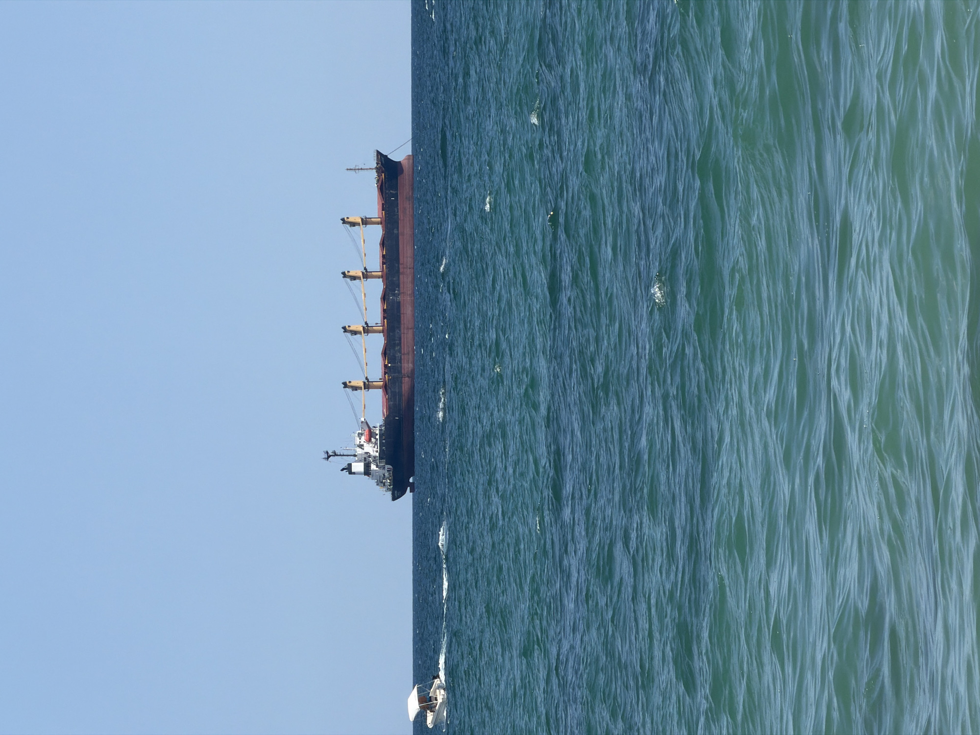 A large cargo ship is visible on the horizon of the Black Sea, with a smaller motorboat in the foreground to the left. The cargo ship is dark red and black with yellow cranes. The motorboat is white with a canopy and appears to be moving. The sea is a deep blue-green with moderate waves. The sky is clear and blue. The scene depicts a maritime setting, likely near Varna, Bulgaria, a major port city on the Black Sea coast.