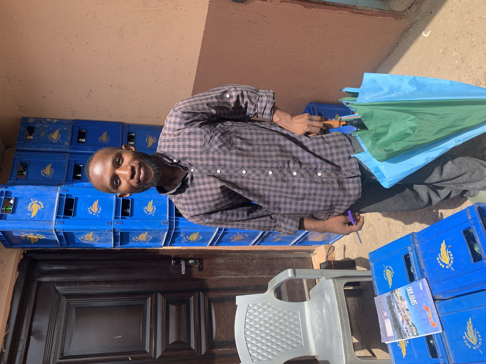 This outdoor daylight photograph captures a man standing amidst commercial crates and other objects, likely in Jos, Nigeria. The man, who appears to be of African descent, has a short beard and dark hair. He is wearing a dark checkered long-sleeved shirt and dark trousers, and is looking directly at the camera with a neutral expression. In his right hand, he holds a purple pen, while his left hand grips the handle of a partially folded umbrella with blue and green panels.
The setting includes a light brown wall in the background and a paved ground surface. To the man's left, a dark brown wooden door with paneling and a silver lock mechanism is visible. A prominent feature of the scene is the numerous blue plastic crates, stacked several high, which bear the text "PREMIUM BREW" and a golden wheat or barley emblem. Some of these crates appear to contain bottles. A light grey plastic chair is positioned near the door, and on top of some crates, a notebook with the title "DREAMS" (and "Vickson" brand) and an image of a coastal town is visible. The strong, direct sunlight indicates it is daytime, likely midday or early afternoon. The overall impression suggests a small business or a shop environment.