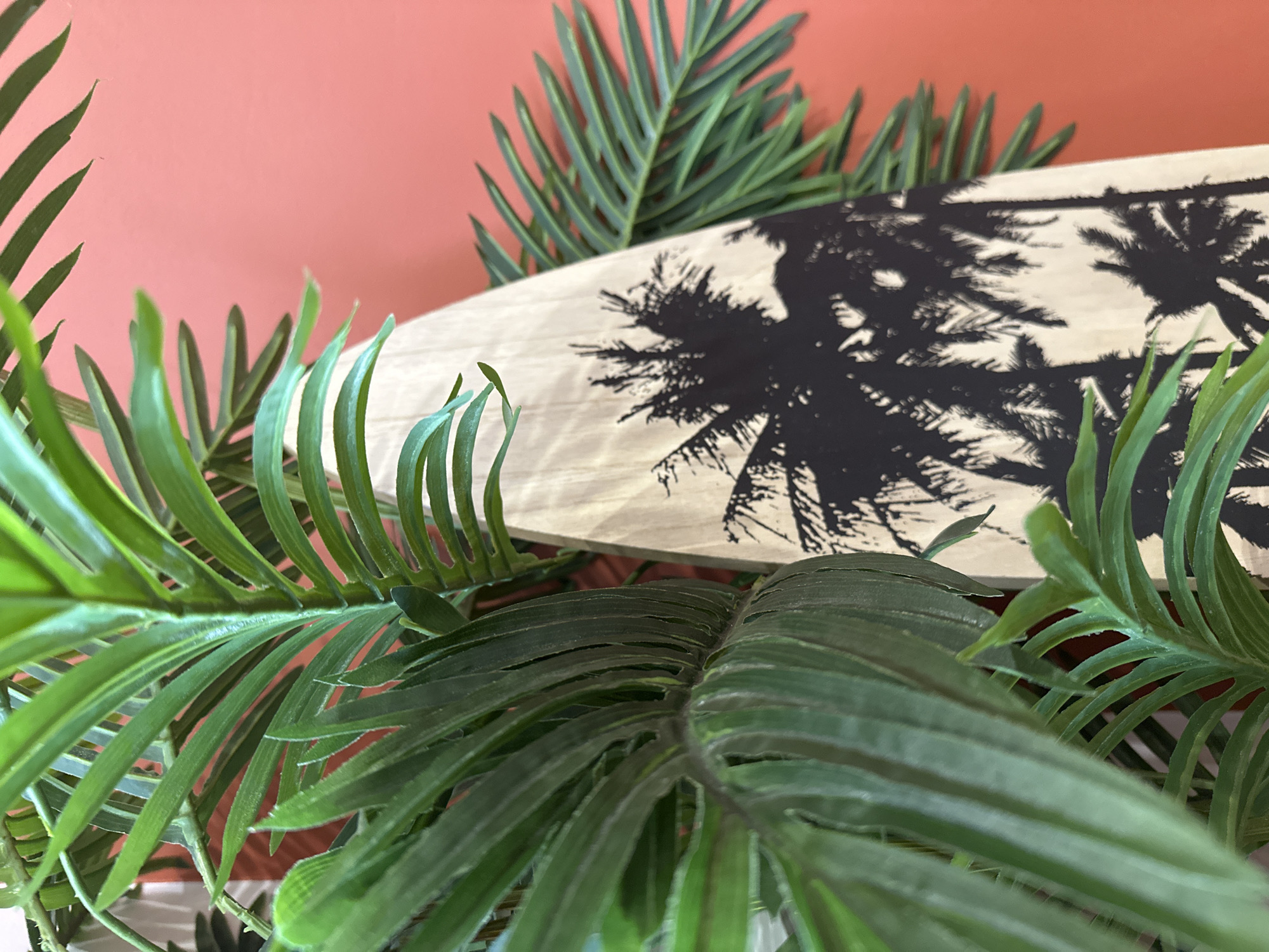 The image presents a decorative scene featuring artificial palm leaves surrounding a surfboard-shaped object with a black-and-white palm tree design. The backdrop is a solid, muted reddish-orange wall. The scene does not appear to be a natural outdoor environment. The composition has an artificial, stylized aesthetic, typical of interior decoration. There is no indication that this image originates from the city of Chaikovsky, Russia.