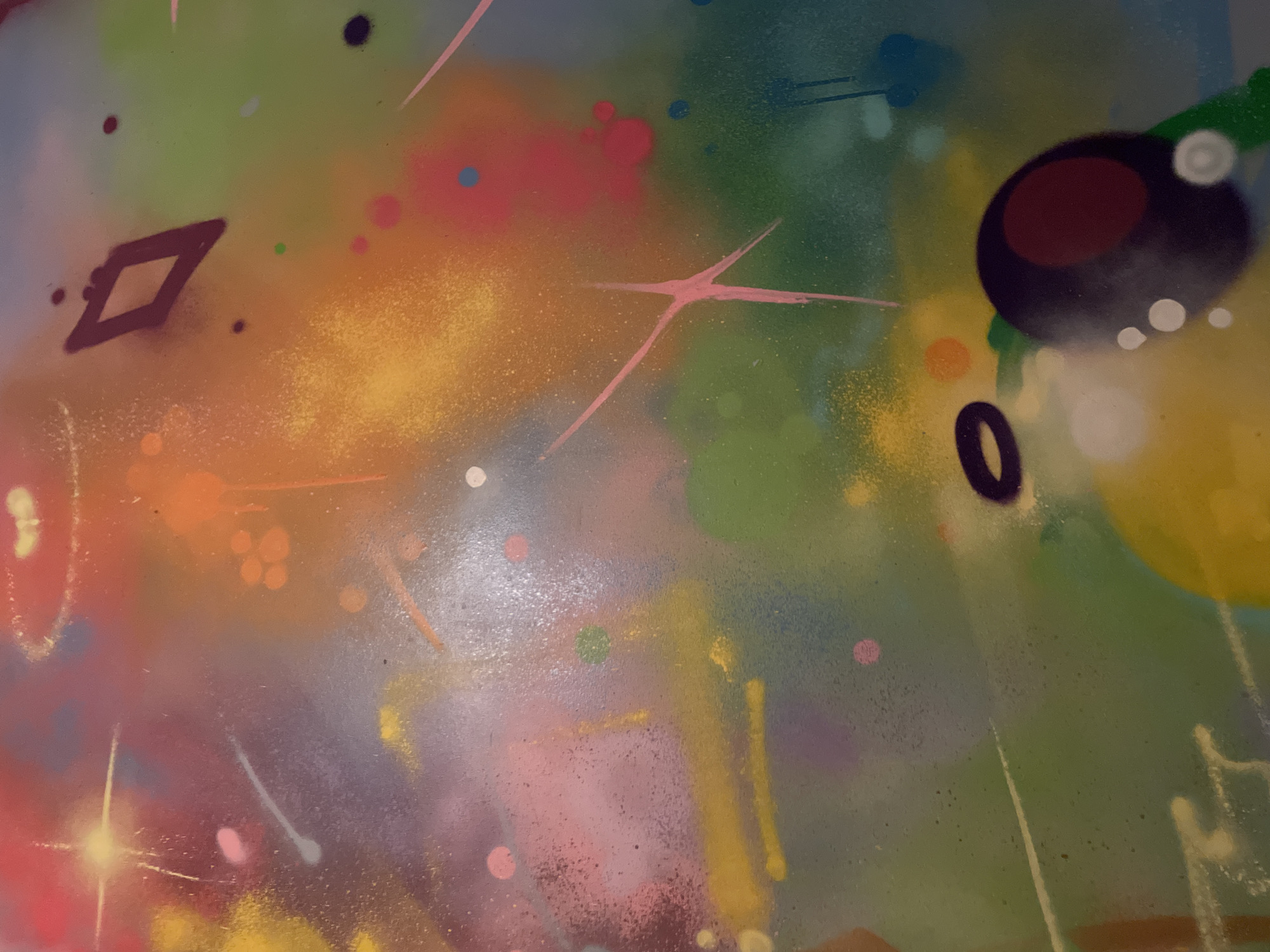 This is a close-up, high-resolution forensic image of an abstract, multi-colored surface, likely a painted wall or canvas, located in the city of Unknown, Unknown. The surface exhibits a vibrant palette dominated by blended tones of orange, yellow, red, pink, green, blue, and purple, applied with techniques suggestive of spray painting and brushwork. Numerous small, irregular paint splatters and larger, diffuse color splotches are visible across the entire frame. Prominent features include a bright, four-pointed star-like burst in the upper-left, several thin, linear streaks (some white, some pink) traversing the composition, and an X-shaped mark formed by two intersecting pink lines in the mid-right. Geometric elements comprise a hollow, dark diamond in the upper-middle, a dark, hollow oval ring in the bottom-middle, and a large, dark circular shape containing a smaller red circle in the bottom-right. Areas of intense yellow and orange provide a luminous effect. The texture of the surface appears slightly irregular, reflecting light inconsistently, indicating a semi-gloss or textured finish. No human figures or discernible actions are depicted; the scene solely focuses on the static painted surface.