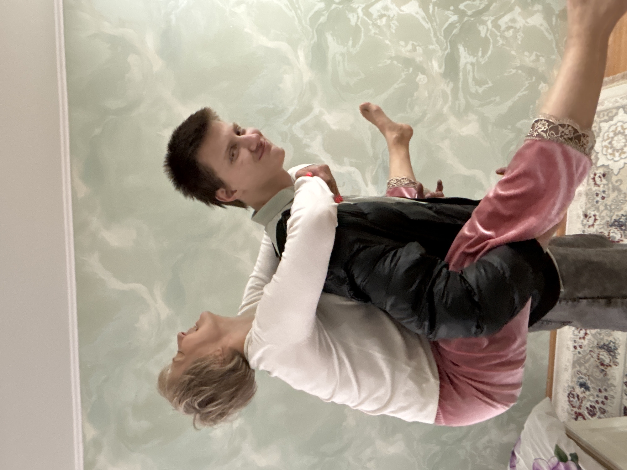 Two people are embracing. One appears to be a young adult male with short brown hair, wearing a white shirt and black jacket. The other appears to be an older female with blonde hair, also wearing a white shirt and pink velvet pants. The background is a wall with a mottled paint effect. A section of patterned rug is also visible. The location is Chaikovsky, Russia. There is no specific action happening beyond the embrace.