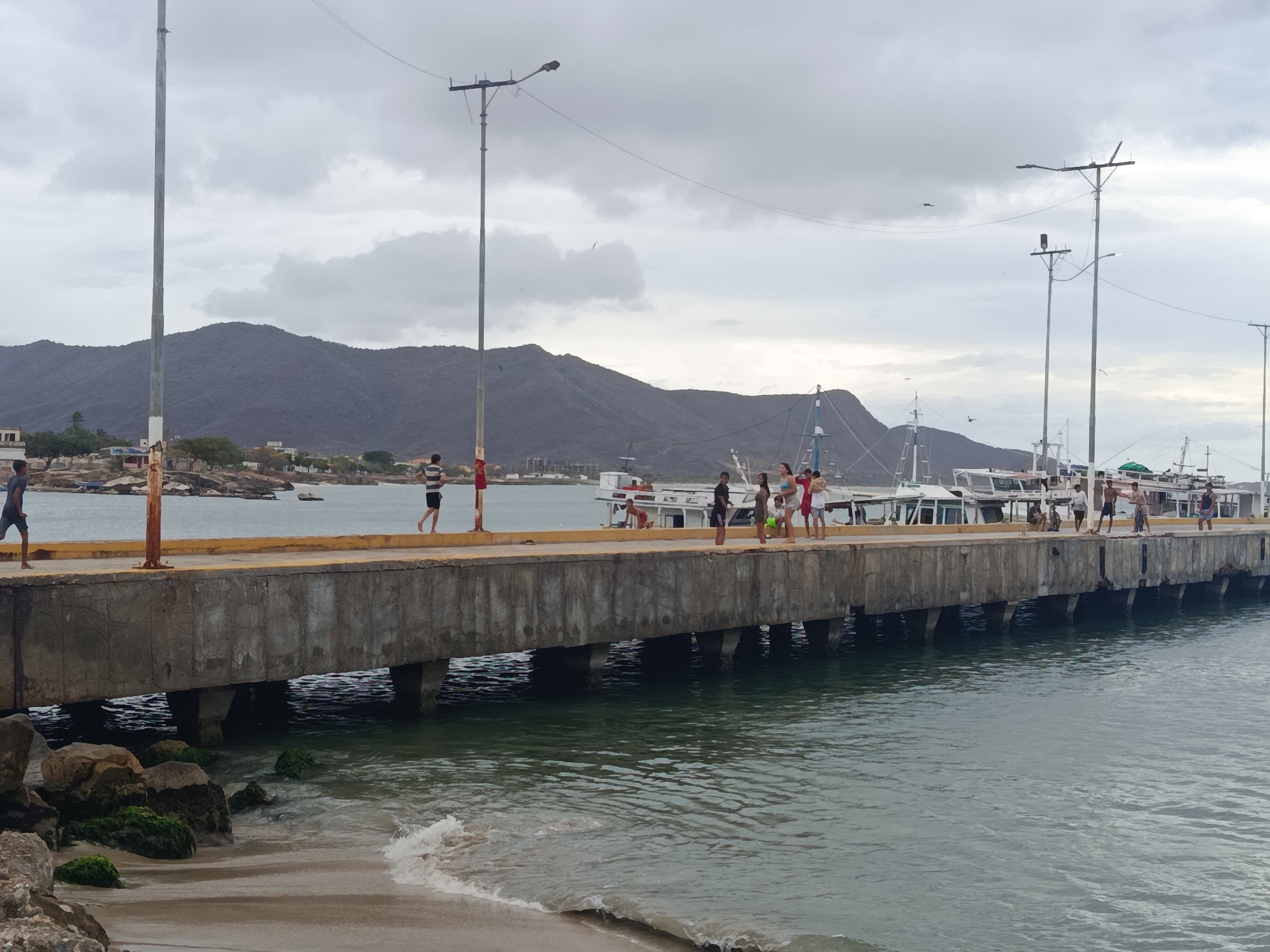 This image captures a coastal scene at a concrete pier in Juan Griego, Isla Margarita, Venezuela, under an overcast sky.
A grey concrete pier extends from the left foreground towards the right mid-ground, supported by multiple concrete pillars submerged in the calm, greenish-blue water. The pier's top edge is marked by a yellow painted strip. Scattered algae-covered rocks and light brown sand form a narrow beach in the immediate foreground, with small waves gently breaking.
Approximately 15-20 individuals are present on and around the pier. On the left side of the pier, a man in a dark t-shirt and shorts walks barefoot towards the left edge of the frame. Further right, another man in a striped shirt and dark shorts walks away from the viewer. A group of individuals, including children and adults in casual attire (shorts, t-shirts, swimwear), are gathered on the pier in the mid-right, some appearing to observe the water. One person is partially visible kneeling or crouching near the pier's edge.
Four tall, grey metal poles with streetlights are positioned along the pier. Electrical lines are visible connecting some of these poles. Several white and blue fishing or passenger boats are docked or moored alongside the pier on the right side of the frame.
In the background, a range of dark, rugged mountains dominates the horizon, with coastal buildings of the town visible at their base. The sky is uniformly grey and heavily overcast, and at least two birds are visible in flight against it. The overall lighting is soft and diffused.