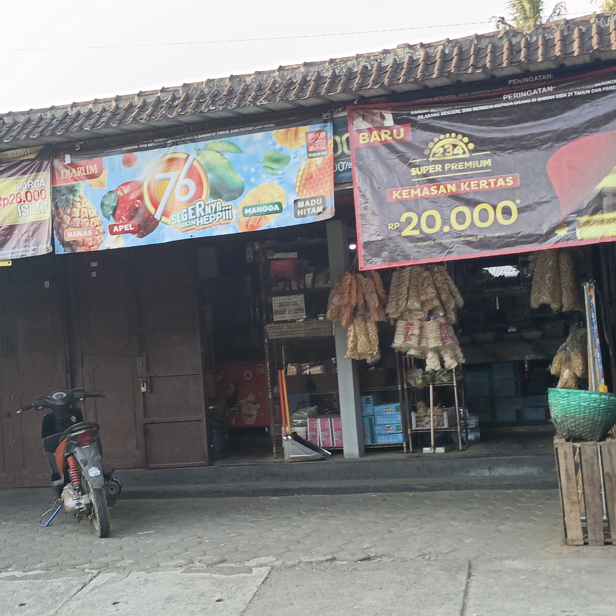 The image captures a street scene in Sraten, Indonesia, featuring the facade of a shop. The shop's roof is tiled, and banners are displayed across its front. To the left, a banner with a yellow background advertises "HARGA Rp28.000 ISI 20" and images of pineapple and apples, along with text like "DIARUM". To the right, a large black banner with red and yellow accents proclaims "BARU SUPER PREMIUM KEMASAN KERTAS RP 20.000*". The shop entrance is a dark brown double door, partially open, revealing shelves stocked with various goods, including drinks and what appear to be snacks. A motorcycle is parked in front of the shop, facing left, with its kickstand down. Beyond the shop, more shelves with products are visible, including a green woven basket on a wooden stand. The ground is paved with cobblestones. The overall impression is that of a small, local business.