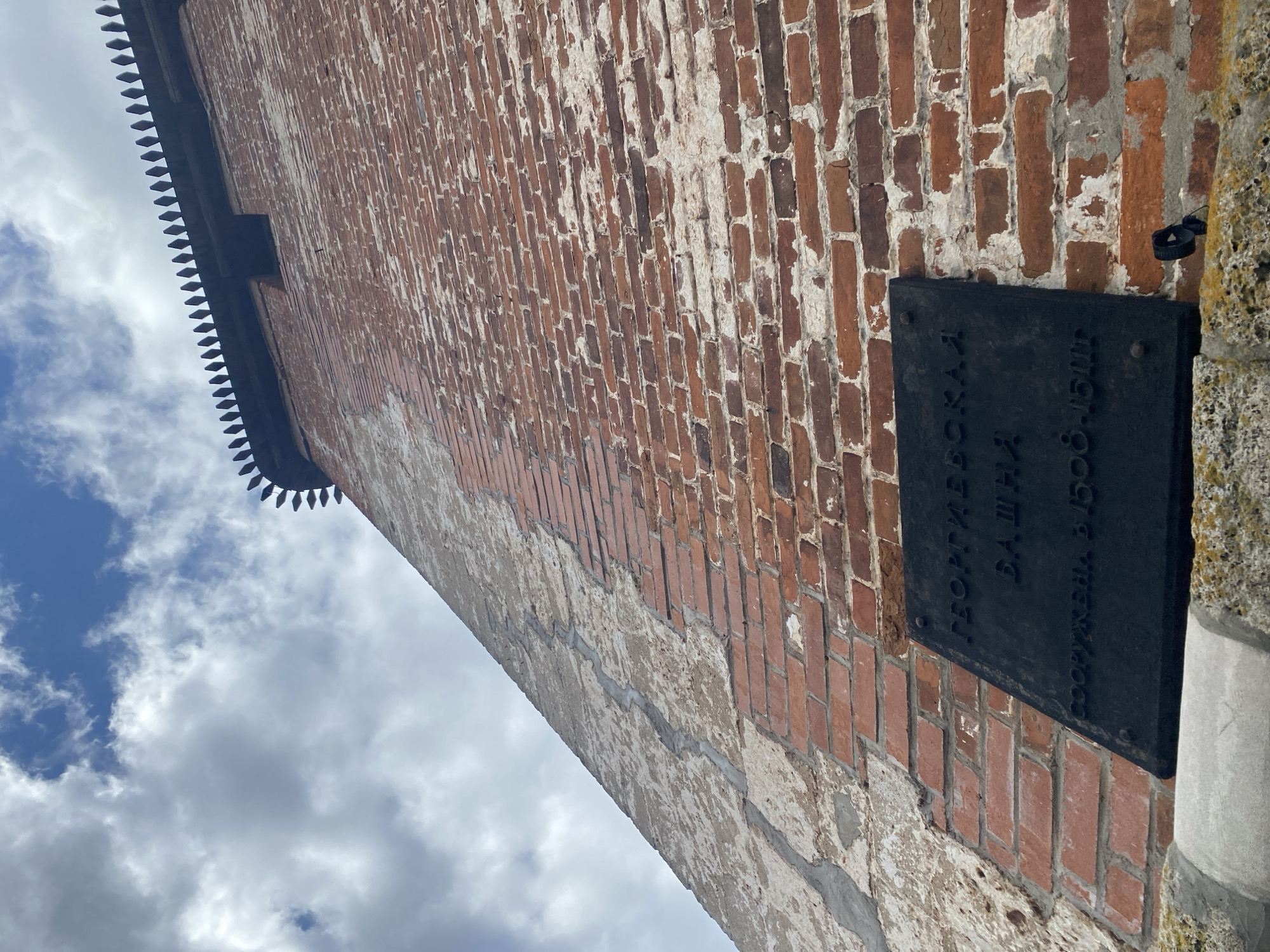 The image presents an upward view of a historic red brick wall, likely part of a fortress or tower. The top edge of the wall features a distinctive crenellated or serrated profile against a bright, partly cloudy sky, indicating daytime.
The wall itself is constructed from reddish-brown bricks, showing signs of age and weathering, with visible mortar and patches of lighter-colored material, possibly remnants of plaster or paint.
A rectangular, dark-colored plaque is affixed to the brickwork, slightly right of the image center. The plaque bears three lines of white Cyrillic text. The first two lines read "ГЕОРГИЕВСКАЯ БАШНЯ," which translates to "George's Tower" or "St. George Tower." The third line, in smaller font, states "СООРУЖЕНА В 1500-1510Г.," meaning "Built in 1500-1510." This identifies the structure as the Georgievskaya Tower, a well-known part of the Nizhny Novgorod Kremlin in Russia, constructed between 1500 and 1510.
To the right of the plaque, a rough-textured, light-colored stone or concrete architectural element is visible. A small, dark, looped fixture is also attached to the wall near this element. The scene is static, focusing on the architectural details and historical plaque.
