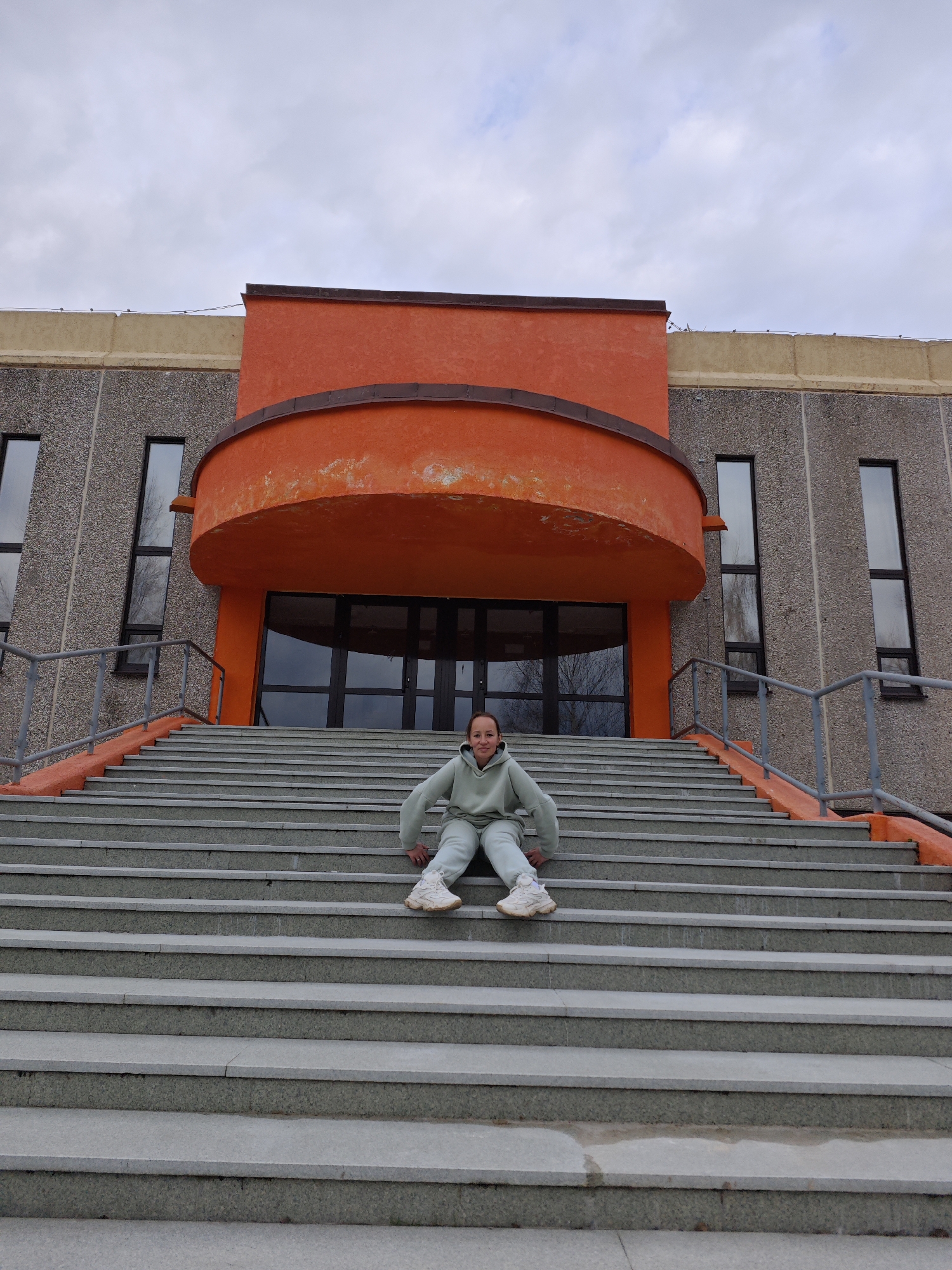A person sits on gray concrete steps in front of a large building, likely in Марковский, Russia. The building has a gray, concrete facade with tall, rectangular windows. An orange, semi-circular structure protrudes from the building, above the doorway. The person is wearing a light-green tracksuit and white sneakers. A gray sky is visible in the background.