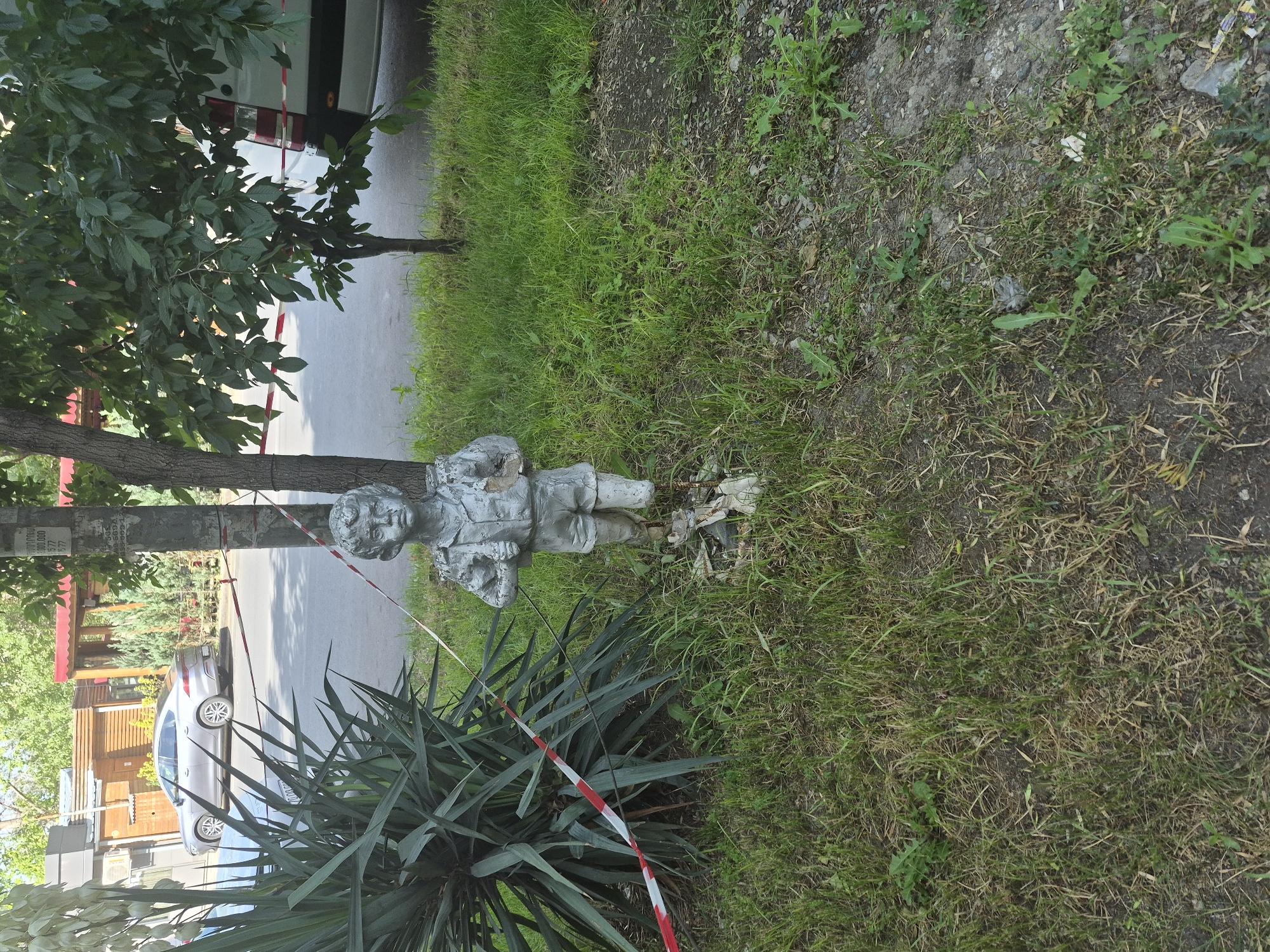 In Yerevan, Armenia, a weathered metal statue of a young boy stands in the grass near a tree and a large yucca plant. The boy is depicted wearing a shirt and shorts, with a backpack on his shoulders, and appears to be walking or standing with his hands held in front of him. Red and white caution tape is draped around the yucca plant and extends towards the statue, suggesting the area might be cordoned off. In the background, a silver sedan is parked on a paved street next to some buildings, and a white van is partially visible. The scene is illuminated by daylight, casting shadows on the ground.