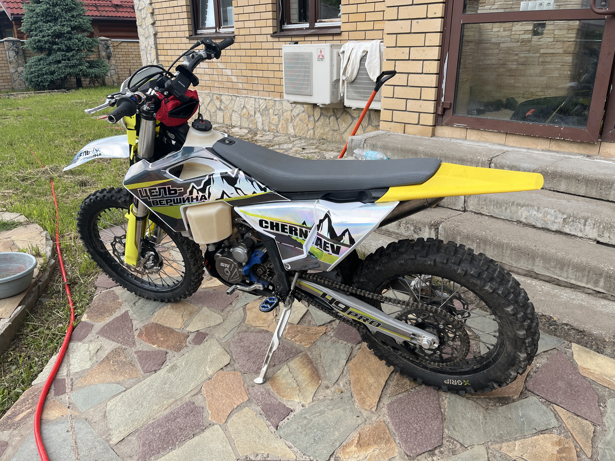 An enduro or dirt motorcycle is prominently featured, parked on a multi-colored flagstone patio next to a house in Oреховка, Russia. The motorcycle is positioned at a slight angle, facing left, with its kickstand down. It features a bright yellow front fender and rear tail, a black textured seat, and translucent fuel tank sections on both sides. Custom graphics in white, black, and light yellow/green adorn the bodywork, including stylized mountain peaks. A red bag is attached to the handlebars. The heavily treaded, knobby tires show signs of dirt, suggesting recent off-road use.
Visible text on the motorcycle includes "ЦЕЛЬ-ВЕРШИНА" (meaning "GOAL-SUMMIT" or "PEAK"), "CHERN AEV", "49PRO" on the swingarm, and "X-GRIP" along with "140/" on the rear tire. The engine casing also shows "SN".
The background includes a house with a brick upper structure and a stone foundation. Two white Mitsubishi Electric air conditioning units are mounted on the brick wall. A large window with a dark frame reflects the surroundings. To the left of the bike, a red garden hose lies on the grass near a grey bucket. A tall green pine tree stands in the distance. An orange-handled shovel leans against the concrete steps leading to the house. The scene is illuminated by daylight.