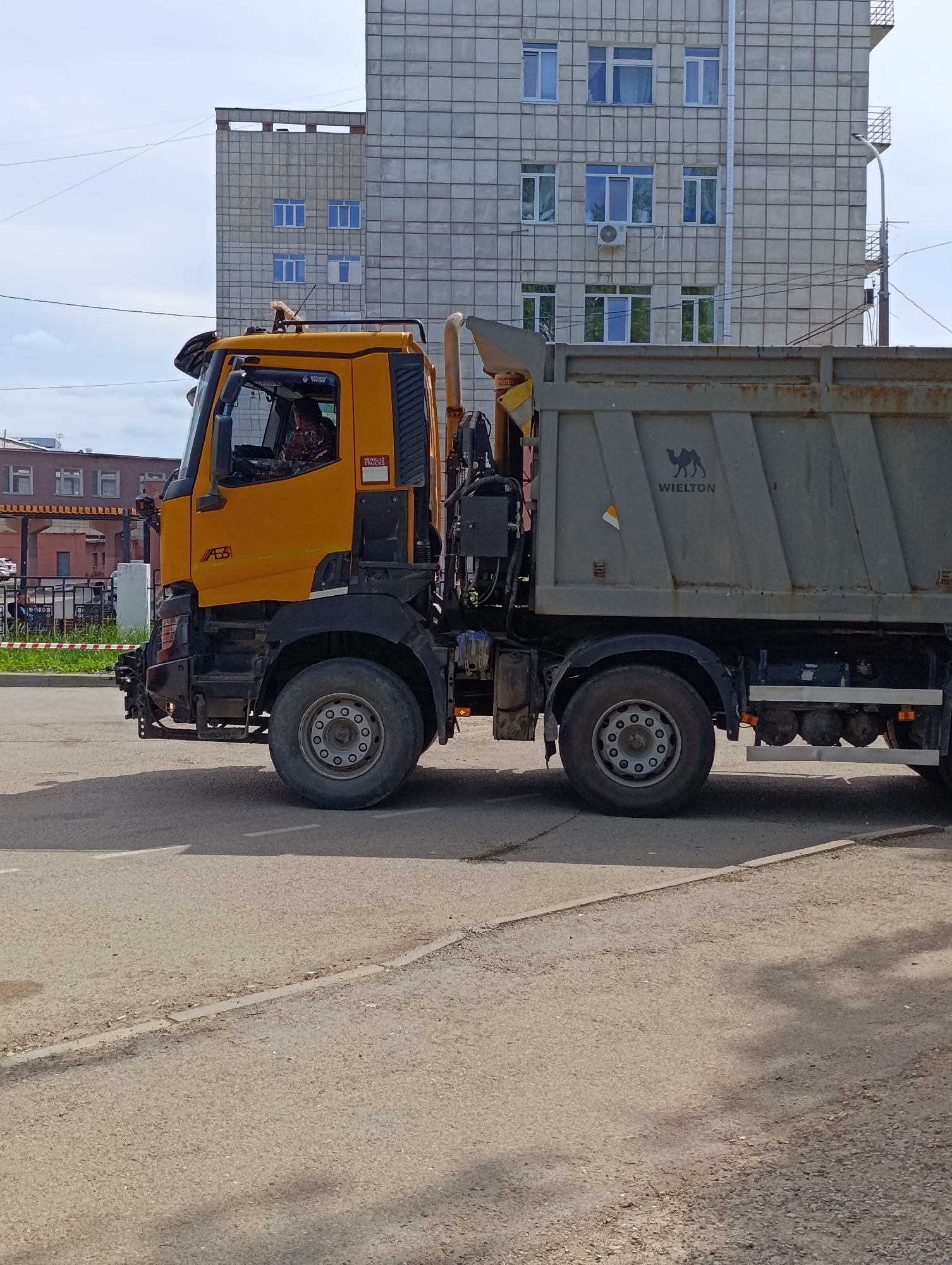 A large, yellow and grey dump truck or specialized vehicle is the primary subject of this daytime image, set in Chaikovsky, Russia. The truck is positioned on an asphalt road, partially obscuring the background. The truck's cab is yellow, featuring the text "RENAULT TRUCKS" and "A63" on its side, along with the Russian acronym "УОКХ" (UOKKh) on a red sticker. A person, presumably the driver, is visible inside the cab. The dump bed is grey, with "WIELTON" and a camel logo stenciled on its side.
In the background, a large, multi-story building with a grey, grid-patterned facade dominates, featuring numerous windows, many with a blue tint and white frames. An air conditioning unit is visible below one of the windows, and a white downspout runs down the side of the building. To the far left, another building with a reddish-brown hue can be seen. The sky above is clear with scattered clouds. The road has faint white lines, and a curb with a narrow strip of green grass and a red-and-white striped barrier are visible to the left of the truck. The overall scene suggests an urban or industrial area.