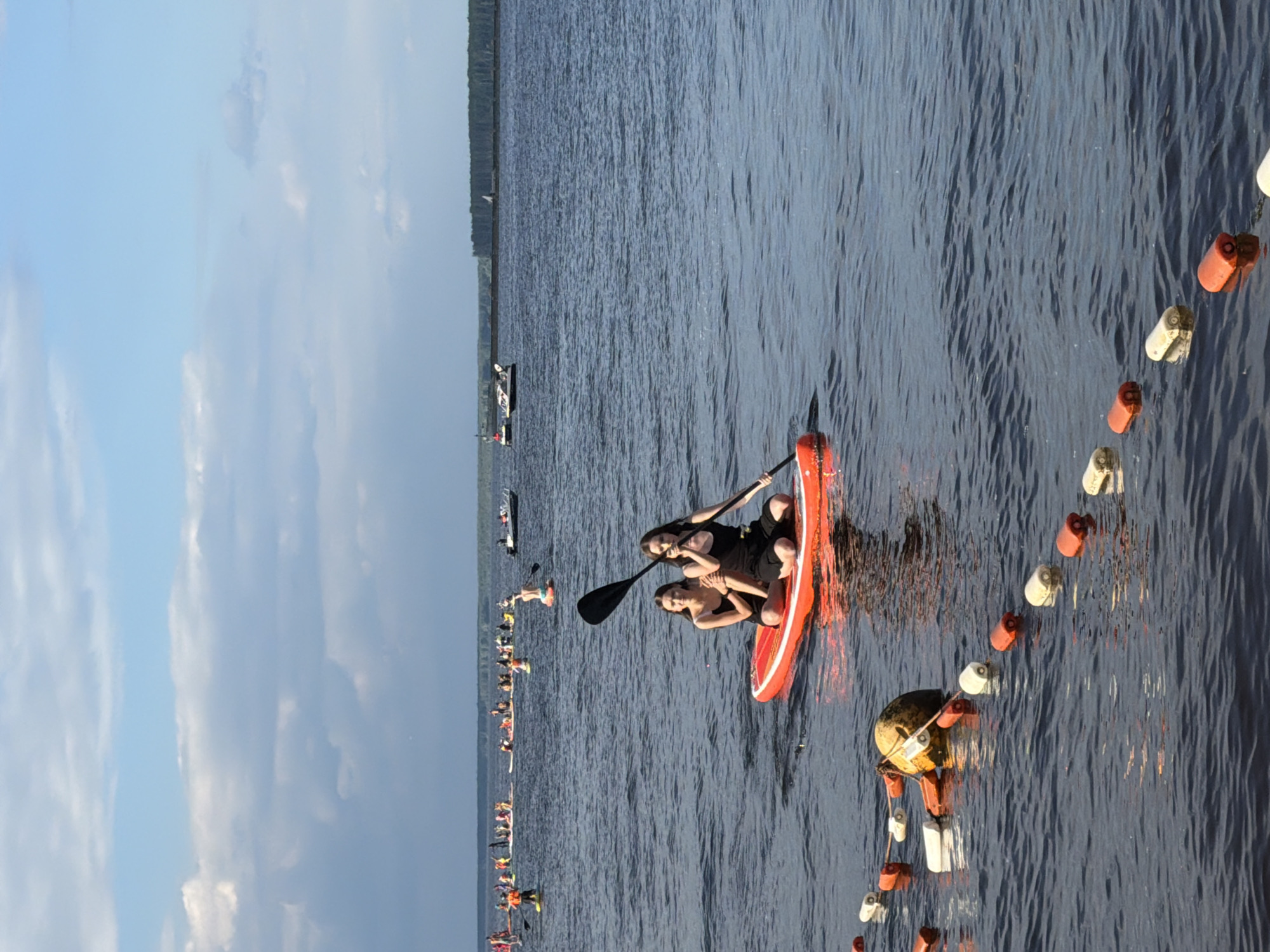 Two women are paddleboarding on a body of water, likely a river or lake, in Chaikovsky, Russia. They are on a single orange paddleboard, with one woman steering and the other seated behind her. Both are wearing black swimwear. In the background, a large group of people on paddleboards are visible, suggesting a recreational event. The water is calm with small ripples. A line of buoys floats in the foreground, marking an area of the water. The sky is partly cloudy with a mix of blue and white. A treeline is visible in the distance on the far shore. The overall scene depicts a summer day of outdoor water activities.