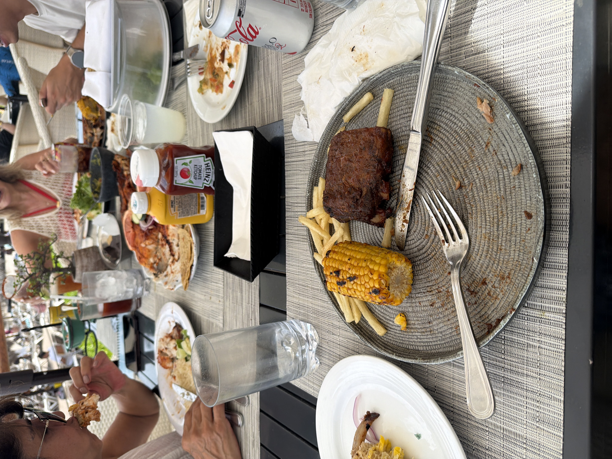 The image depicts a table setting in what appears to be an outdoor dining area. The scene includes remnants of a meal, suggesting that people have recently been eating at the table. The table is covered with a textured, possibly woven, placemat.
Present are multiple plates, some empty or containing food scraps. One plate has leftover ribs, french fries, and corn on the cob. There are also visible condiments, including ketchup and mustard bottles. Used napkins and empty beverage containers (e.g., a Coca-Cola can) are also present on the table.
Several people are visible, with some captured in the act of eating. One person is eating a piece of food with their hand. Other people are seated at the table, some partially obscured.
The setting appears to be outdoors. The location in the city of Las Jarretaderas, Mexico, is not directly depicted in the photo, therefore, this detail cannot be confirmed from this image.