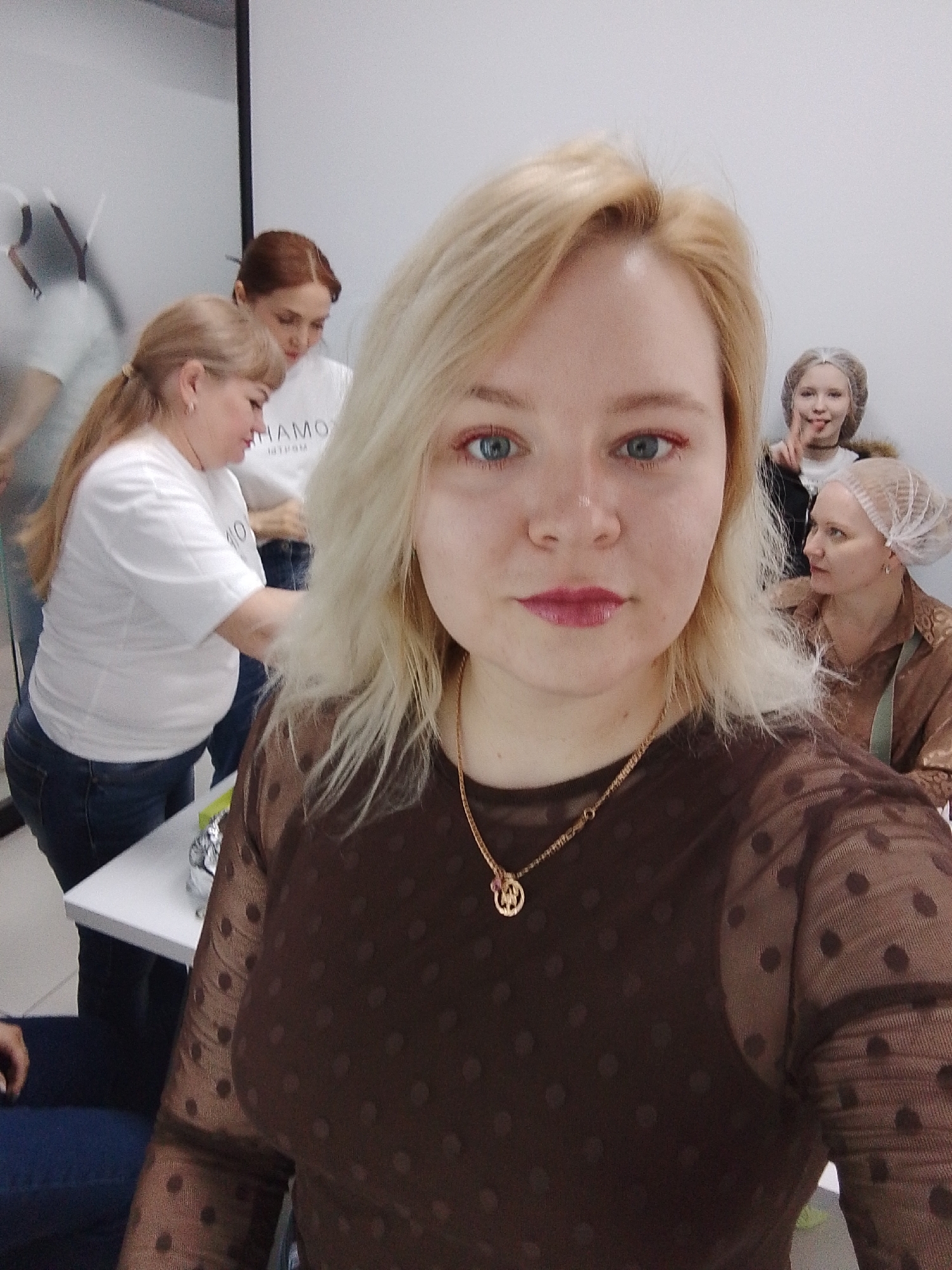 A medium indoor shot depicts a fair-skinned woman with blonde hair, a gold necklace, and a brown polka-dot mesh top, taking a selfie. Behind her, four other women are visible: one with red hair in a white shirt, another with blonde hair in a white shirt tying an apron around the red-haired woman, and two women in hairnets. The location is in Izhevsk, Russia. The environment appears to be a beauty salon or cosmetic studio due to the presence of multiple people getting ready, possible make-up applications, and the beauty-related atmosphere.