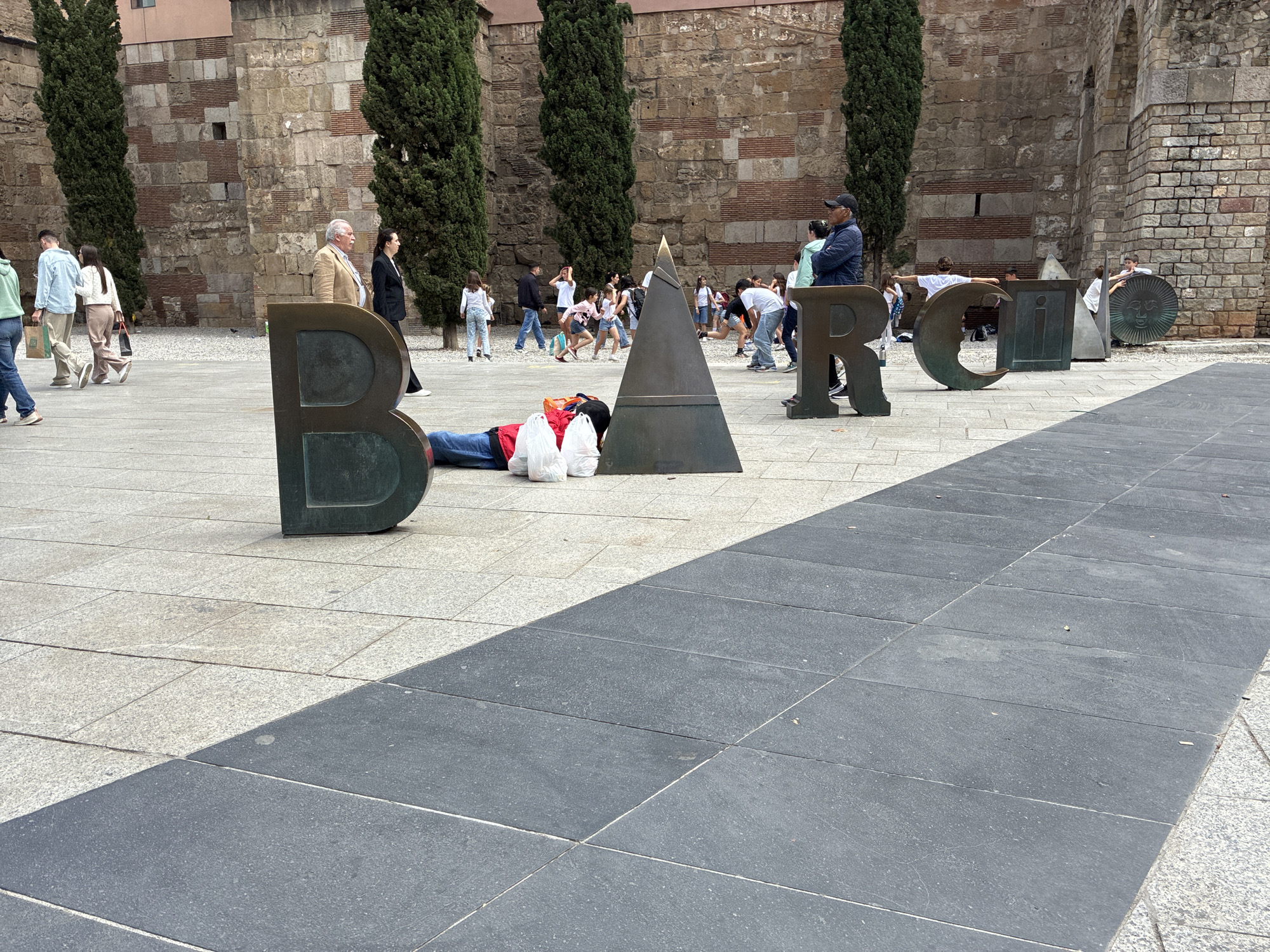 The image shows a public square in Barcelona, Spain, with large metal letters spelling out "BARCELONA". A person is lying down between the "B" and the triangular shaped "A" of the word, appearing to be resting or sleeping. The person is wearing jeans and a red jacket, and has two white plastic bags beside them.
Around the lettering, there are other people walking, standing, and taking photos. Some are walking towards the viewer, while others are gathered near the end of the sculpture. In the background, there is an old stone wall with brick sections, tall, thin trees, and parts of buildings. The foreground of the square is paved with light-colored stones, contrasted by an area of darker gray stone tiles.