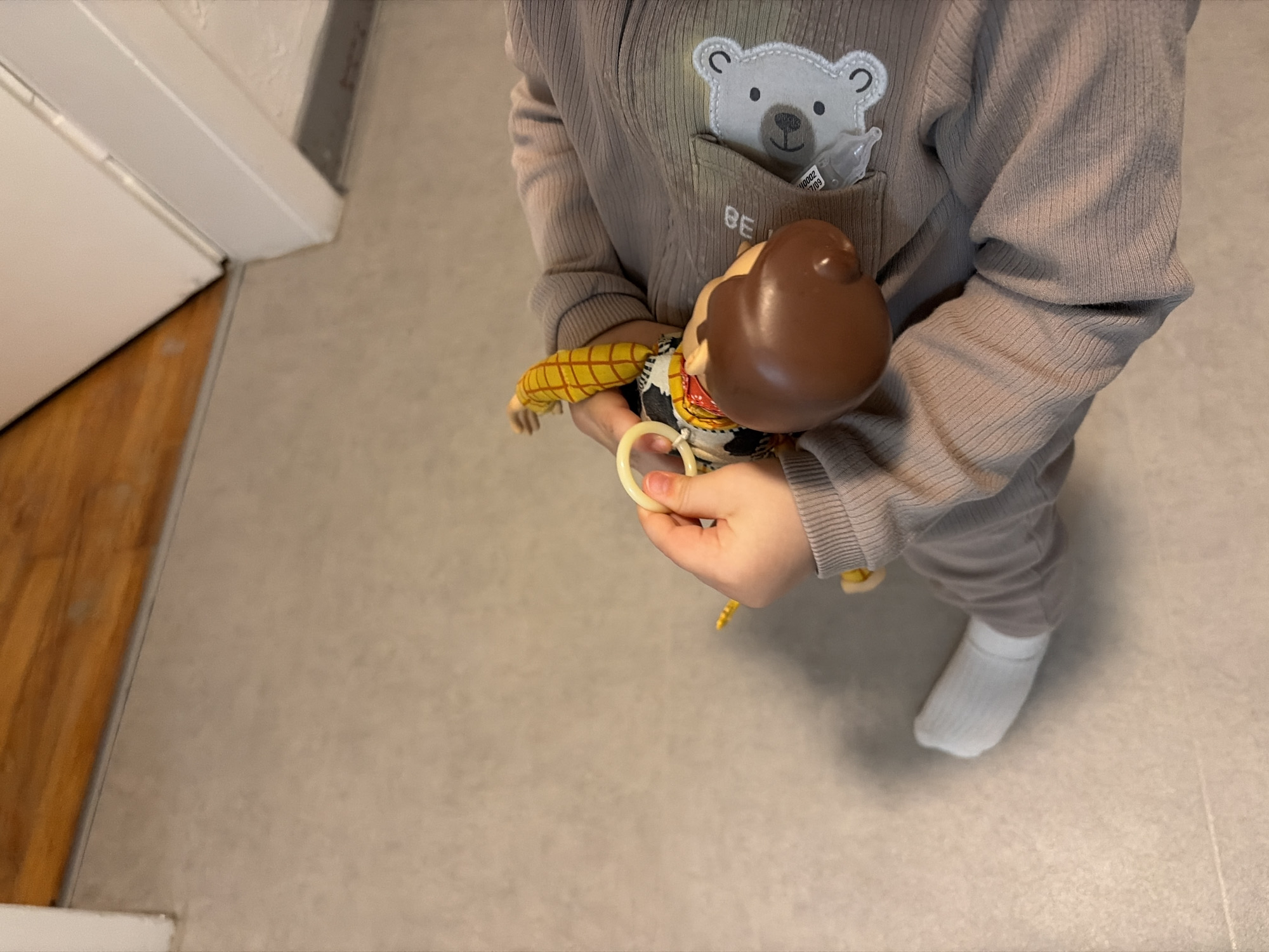 A child, dressed in a gray, corduroy-like jumpsuit with a bear design on the chest pocket, is holding a Woody doll. The child is also wearing a white sock. The scene is set indoors on a gray tiled floor next to a white wall and doorframe. The image does not provide sufficient information to place the location in the city of Pétange, Luxembourg.