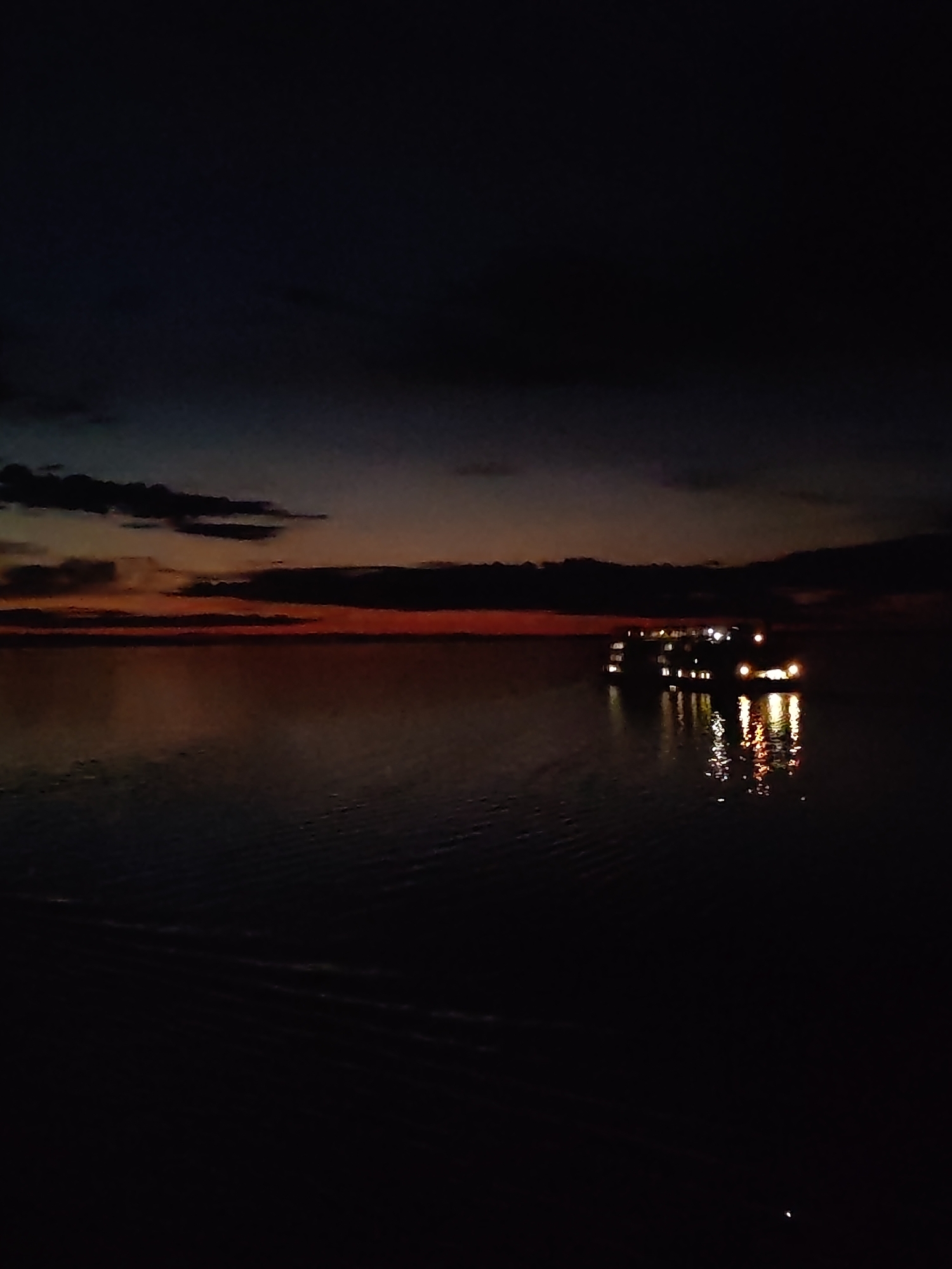 The image captures a nighttime scene at the former location of Mologa, Russia, now submerged by the Rybinsk Reservoir. A large vessel, likely a passenger or tourist boat, is illuminated with multiple lights, reflecting in the dark water. The sky is dark, with a gradient of orange and red at the horizon, indicating either a sunset or sunrise. A few dark clouds are present near the horizon. The overall environment suggests a still, open body of water.
