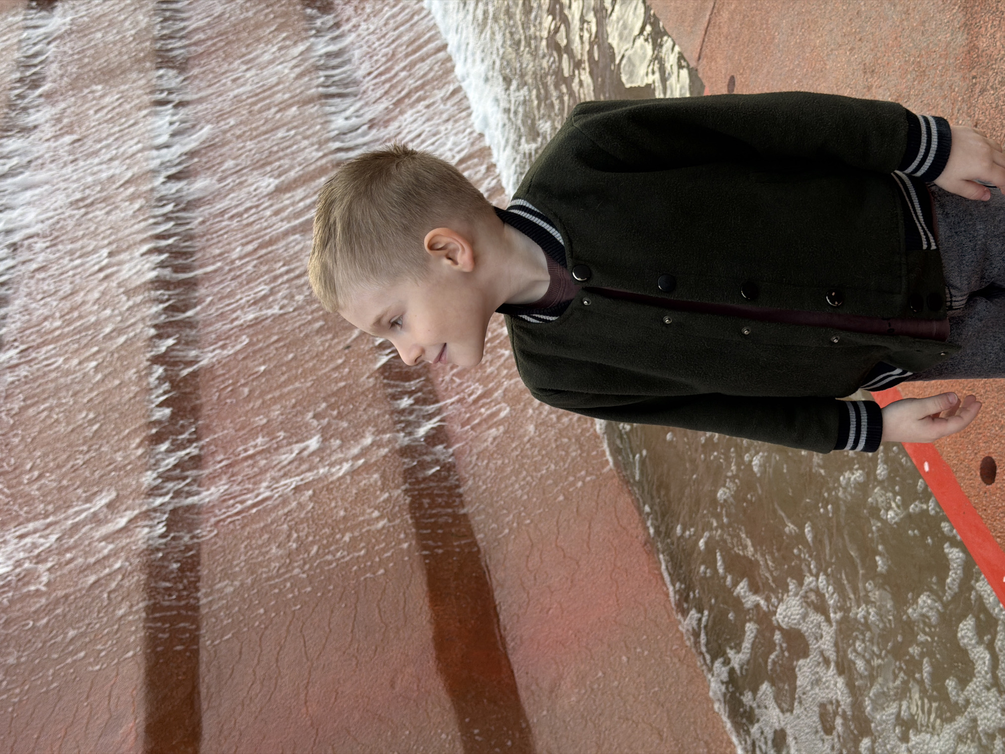 A young boy stands beside a cascade of water on what appears to be an artificial waterfall or decorative water feature. The water flows over a series of steps or ledges, creating a rippling effect. The boy is wearing a dark green jacket with striped cuffs and a button-down front. He looks down at the water. The location is Krasnodar, Russia. The environment appears to be a public area, possibly a park or city square.