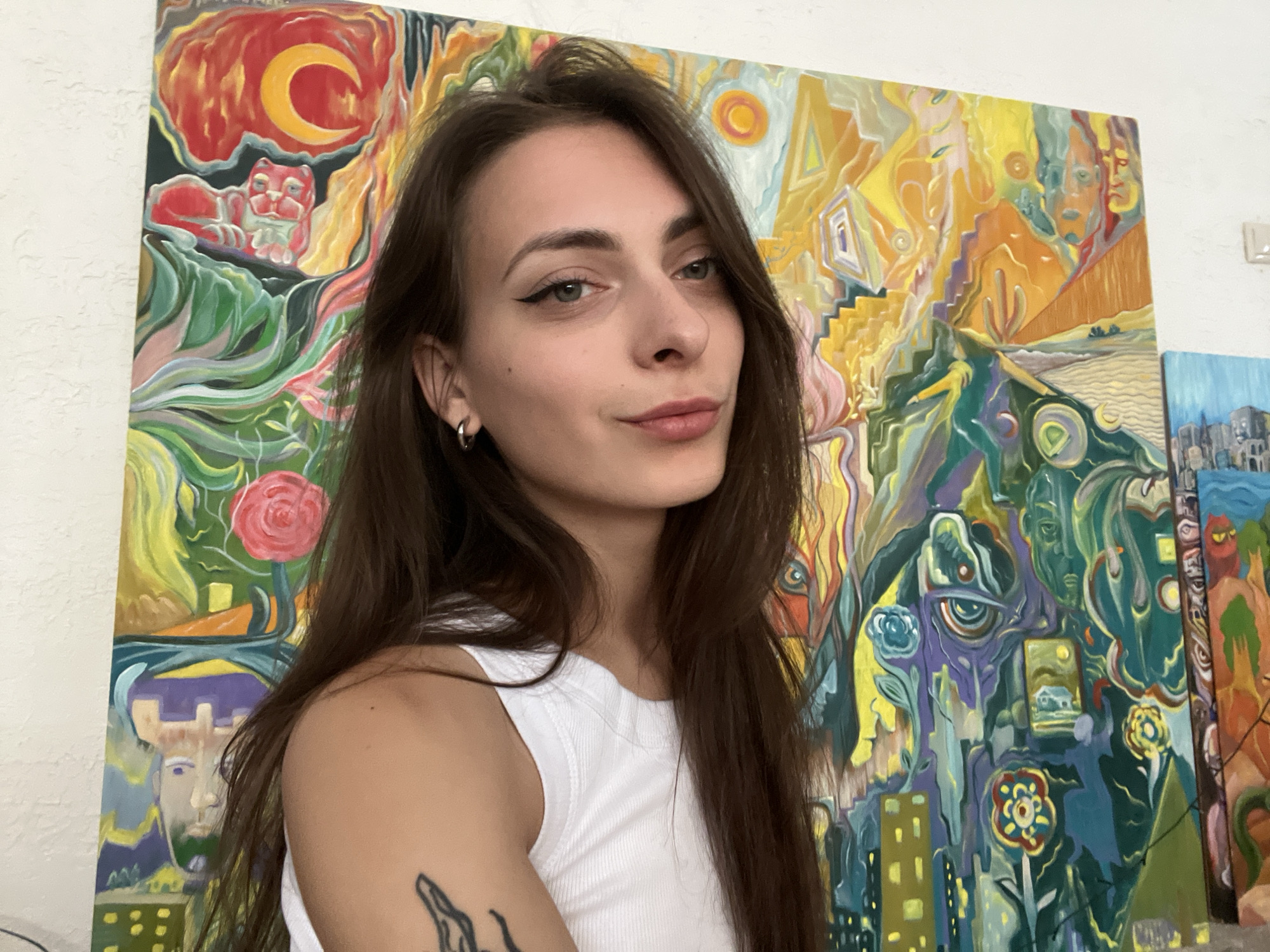 A young Caucasian woman with long, dark brown hair and a white tank top is positioned in the foreground, facing the camera and slightly to the left. She has delicate features, a light complexion, and is wearing eyeliner. A silver hoop earring is visible in her left ear, and she has a tattoo on her left arm. Behind her, a large, colorful abstract painting dominates the background. The painting features a vibrant palette of oranges, yellows, purples, greens, and blues, with stylized figures, surreal landscapes, and what appear to be architectural elements. To the right of the main painting, another smaller painting with a cityscape and a blue sky is partially visible. The overall setting appears to be an indoor space, possibly an art studio or gallery. There is no discernible information within the image to indicate the location as Krasnoyarsk, Russia.