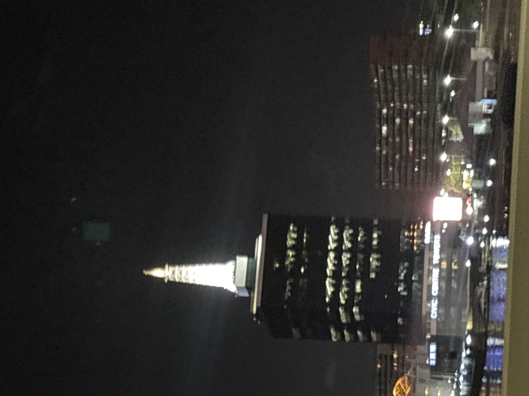 The image shows a nighttime cityscape in Ikoyi, Nigeria. The most prominent structure is the Civic Centre Towers, illuminated with a bright, spire-like top. Other buildings are visible nearby, also lit up at night. The overall environment appears dark, with artificial lighting being the primary source of illumination. The photo seems to have been taken from an elevated position, possibly from another building or a high floor.