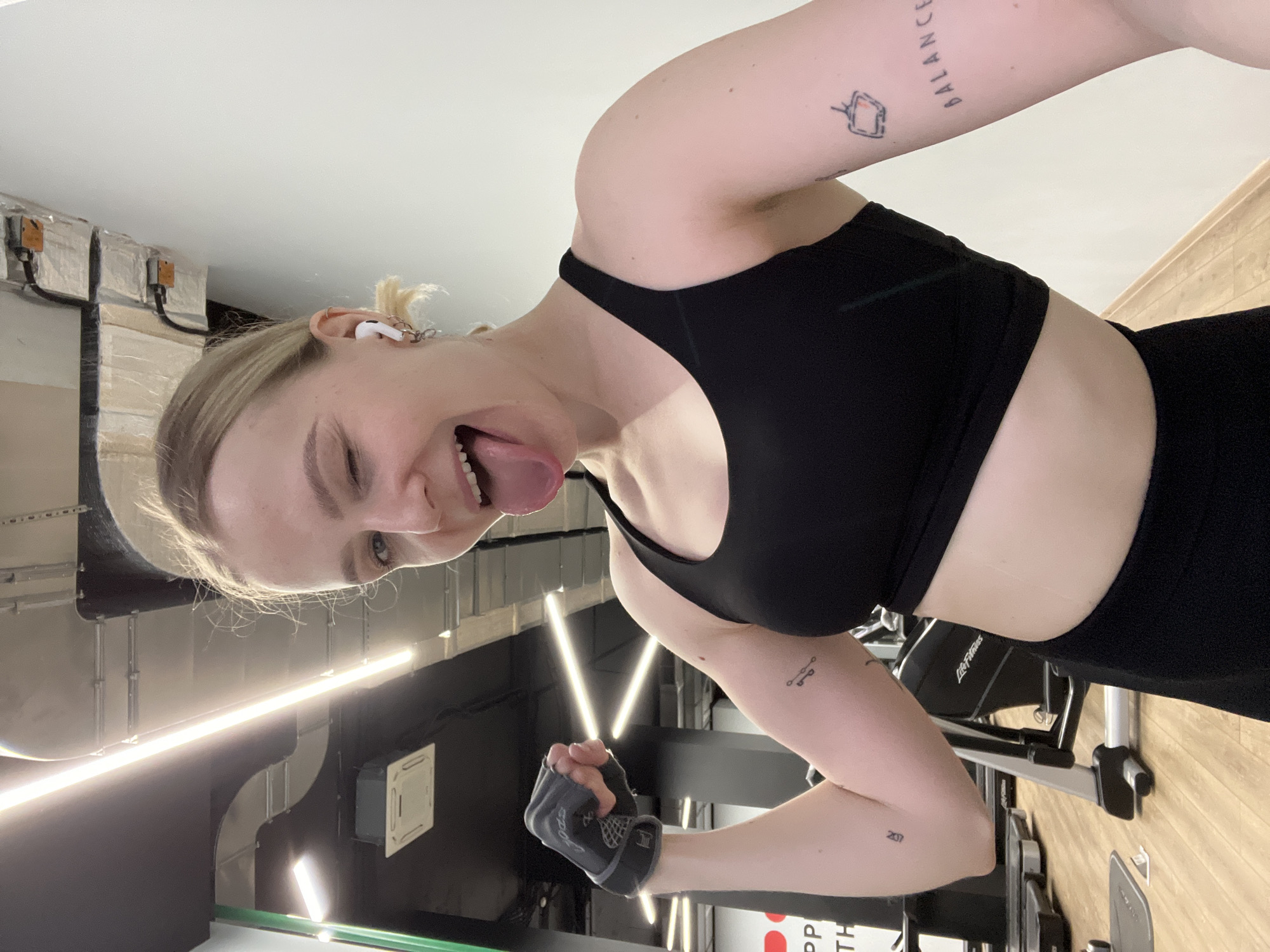 A young woman with blond hair pulled back in a ponytail is posing in a gym. She is wearing a black sports bra and black bottoms. She has an earbud in her left ear and is sticking her tongue out while winking. Her left arm is bent at the elbow, and she is wearing a gray workout glove on her left hand. Tattoos are visible on both arms. The gym has weight machines and bright lights. A sign with Cyrillic characters is partially visible. The gym is located in Khimki, Russia.