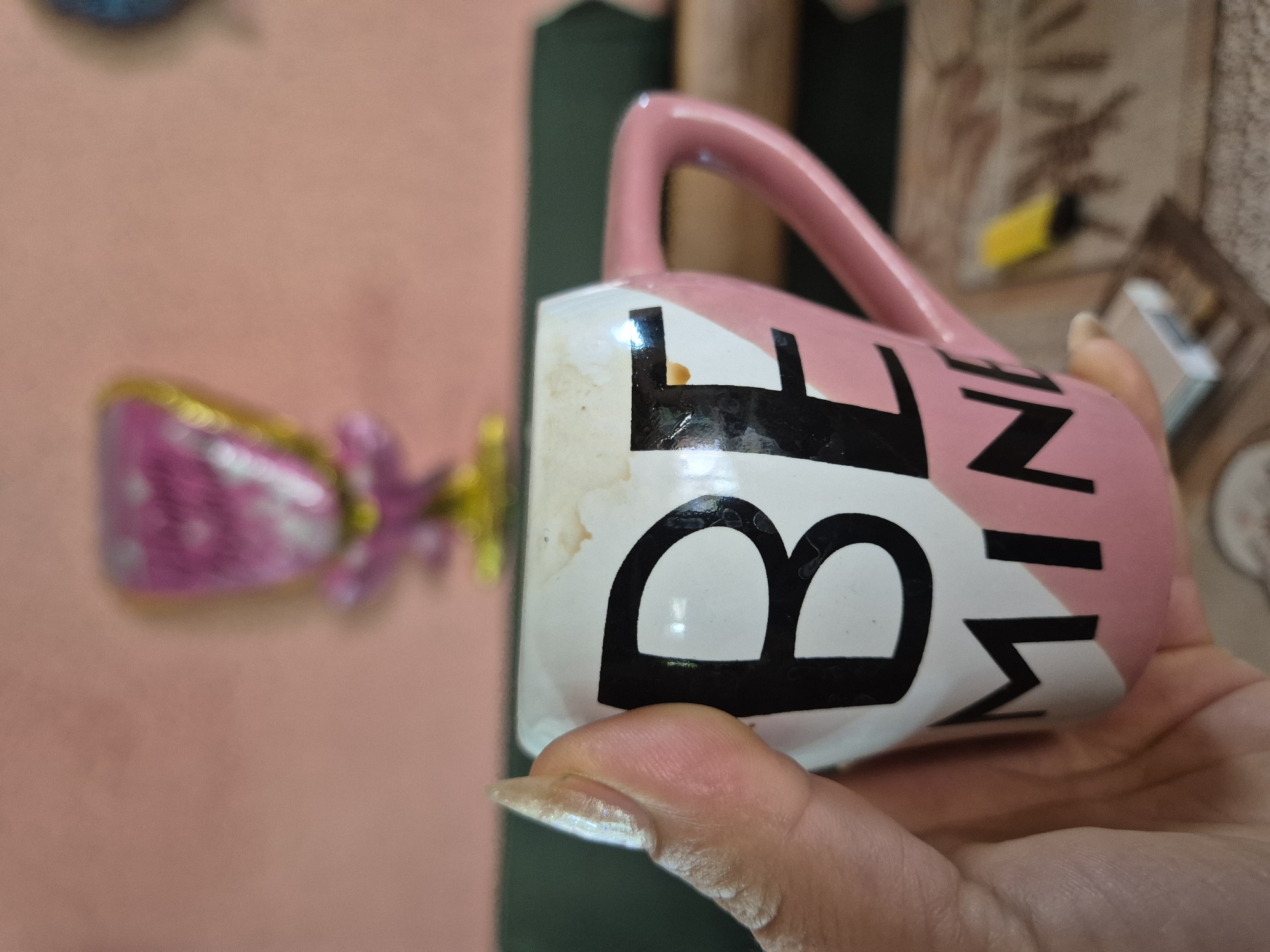 A hand with long, light-colored fingernails holds a pink and white mug with black lettering. The mug has a pink handle and is partially filled with a brown liquid, possibly coffee. The lettering on the mug reads "BE MINE". In the background, a pink wall is visible, with a blurry, heart-shaped balloon and some yellow object. The overall scene appears to be an indoor setting.