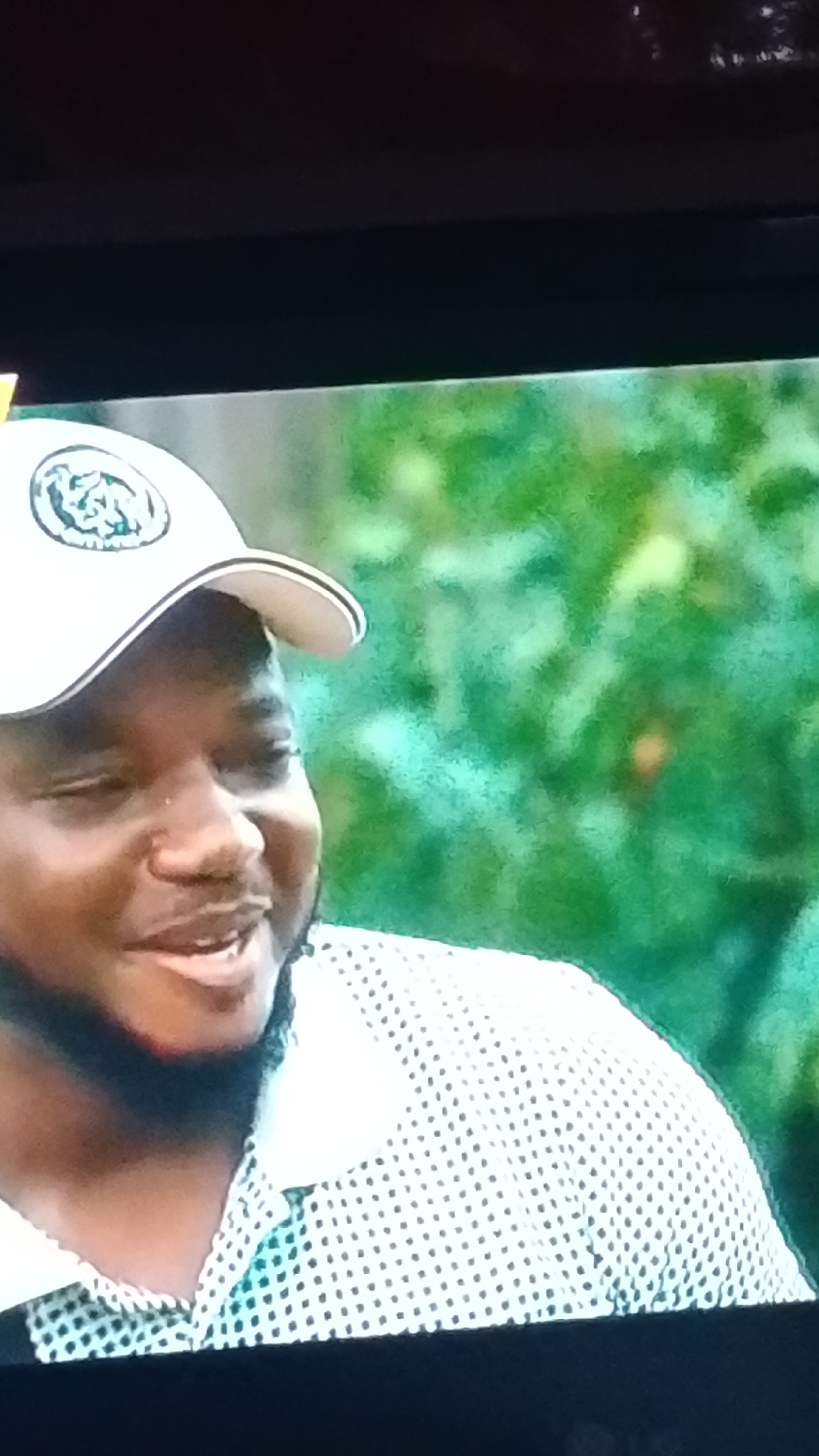 The image depicts a close-up, chest-up shot of an adult male, originating from a screen capture. The scene is located in Lokoja, Nigeria.
**People:**
One male individual is visible. He has dark skin, a short, neatly trimmed beard and mustache, and appears to be of African descent. His expression is a clear, warm smile, with his eyes squinted slightly and his mouth open, revealing some teeth. A small, subtle stud piercing is visible on his left nostril.
**Subjects and Objects:**
The man is wearing a white baseball cap with a contrasting dark trim on the brim. The front of the cap features a circular emblem with an intricate, dark-colored design or logo. He is also wearing a collared shirt, light-colored (appears white or light blue) with a pattern of small, dark, uniform polka dots.
**Actions and Interactions:**
The man is smiling and looking slightly off-camera to his right, suggesting he is reacting to someone or something outside the frame. No direct interaction is depicted within the cropped view.
**Environment:**
The background consists of out-of-focus green foliage, indicative of an outdoor setting such as a garden, park, or natural landscape. The blurriness of the background keeps the focus on the individual. The image itself shows black bars at the top and bottom, consistent with a photograph taken of a television or computer screen.