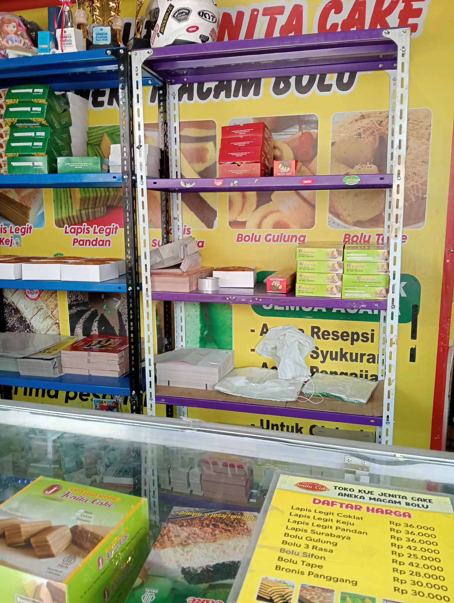 The image shows a display shelf in a cake shop in Grabag, Indonesia. The shelves are filled with various cakes and cake-related items. On the top shelf, there are boxes of "Lapis Legit" cake and "Bolu Gulung" cake. There are also some empty cake boxes and packaging materials. The middle shelf has more boxes of "Lapis Legit" and "Bolu Tule" cakes, along with some promotional materials and a roll of plastic wrap. The bottom shelf displays more cake boxes and packaging. In the foreground, a glass display case holds more cakes and a price list for "Jenita Cake". The shop appears to be well-stocked and organized, with a focus on traditional Indonesian cakes. The overall impression is of a busy and successful bakery.