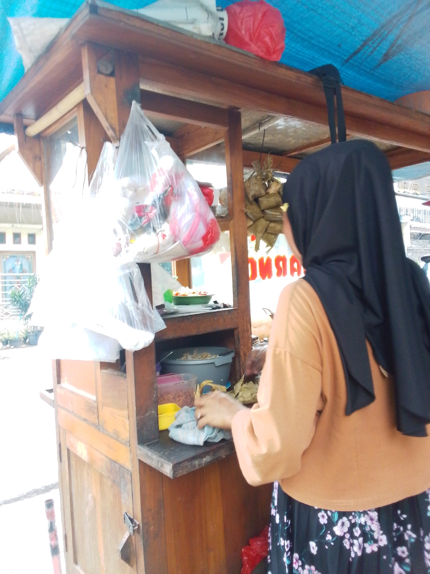 The image shows a street food stall in Central Jakarta, Indonesia. A woman wearing a black hijab and an orange shirt is standing behind the stall, preparing food. The stall is made of wood and has a blue tarp covering the top. There are several plastic bags filled with food hanging from the stall, as well as some wrapped food items. In the background, there is a building with windows. The scene appears to be outdoors, with natural lighting.