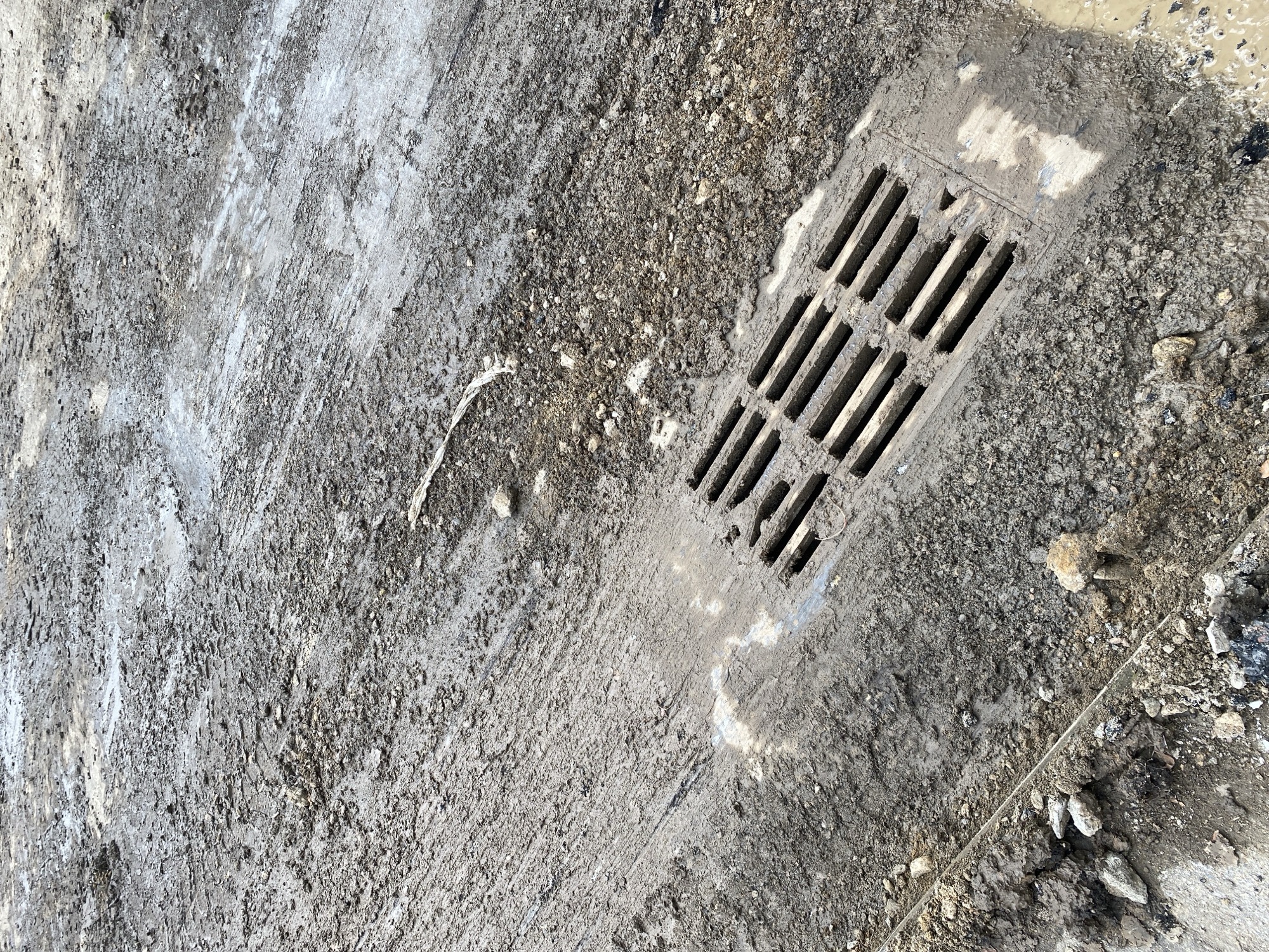 The image shows a close-up, eye-level, outdoor, overhead shot of a muddy, wet, dirt and gravel road. The foreground features a metal storm drain grate, partially covered in mud and debris. Tire tracks are visible in the mud, indicating vehicular activity. The ground is uneven and appears to be undergoing construction or is in a state of disrepair. A small puddle of water is present near the grate. The lighting suggests an overcast day. The location is unknown.