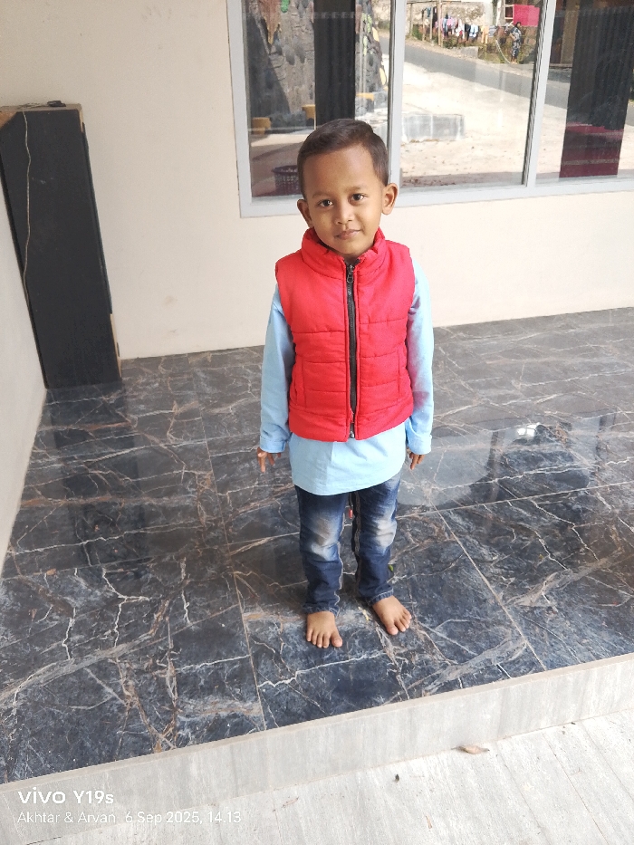 A young child, approximately 3-6 years old, stands centered and facing forward on a dark grey marble-patterned tiled floor with white and brown veining. The child wears a red puffer vest with a black zipper over a light blue long-sleeved shirt and blue denim jeans; the child is barefoot. To the left, a tall, black, rectangular object resembling a speaker stands against a light beige wall. To the right, a multi-pane window in a light frame reveals an outdoor environment featuring a dirt or paved road, distant residential structures, laundry hanging on a line, and sparse vegetation under natural daylight. The image depicts a covered outdoor or semi-enclosed area, likely a porch or patio, located in Sukadana, Indonesia. A lighter grey concrete step or threshold is visible at the bottom of the frame.
