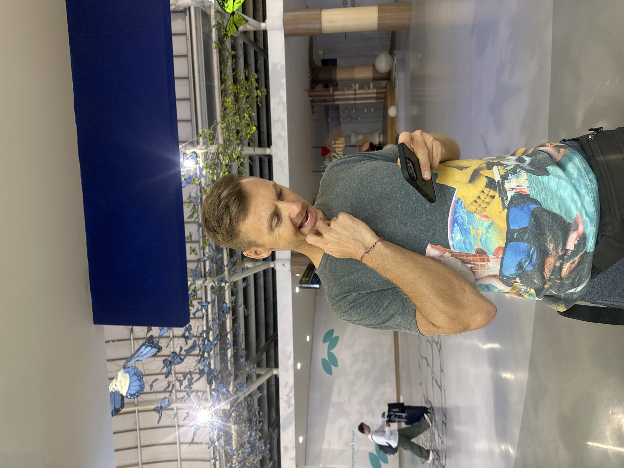The image depicts a fair-skinned man standing in what appears to be an indoor public space, likely an airport terminal, given the context of Kuta, Indonesia. He is positioned slightly to the left of the frame, facing right, and looking towards his black smartphone held in his left hand. His right hand is near his chin/mouth, and his mouth is slightly open, suggesting he might be speaking or contemplating something. He wears a grey short-sleeved t-shirt with a vibrant, multi-colored graphic print featuring various scenes and elements, and grey sweatpants. A red and white braided bracelet adorns his right wrist.
The background reveals a spacious environment with high ceilings and polished light-colored floors. To the left, a large, dark blue fabric panel or screen stands vertically. Decorative blue butterfly cutouts are suspended from the ceiling in the mid-ground, creating an artistic display, alongside some green artificial foliage. Bright artificial lights illuminate the space. Further in the background, another person is seen walking away, pulling luggage. On a distant wall, faint green leaf-like logos and part of the Indonesian text "Seperuh Hasi" (likely "Sepenuh Hati" meaning "wholeheartedly") are visible. A small heart shape and the word "Bali" are also faintly discernible on a sign, reinforcing the location context. The setting suggests a modern, well-lit, and decorated area within a transit hub or large public building.