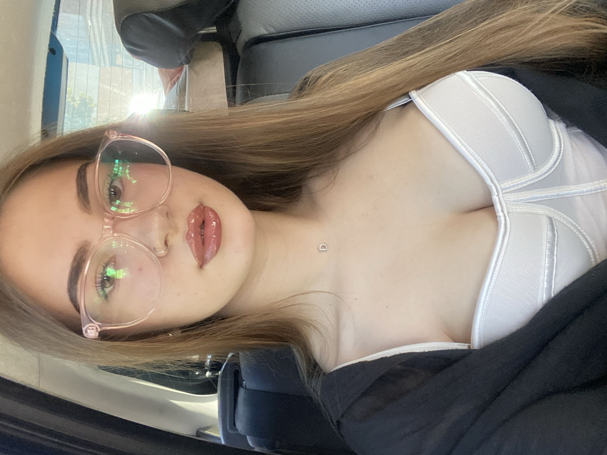 A young woman with long, light brown hair and glasses is pictured in the backseat of a car. She is wearing a white bustier and a black cover-up. A delicate silver necklace with a diamond pendant is visible. The background shows the interior of a car, with a window reflecting sunlight. The scene is not located in the city of Дьяково, Russia; it appears to be a typical car interior setting.