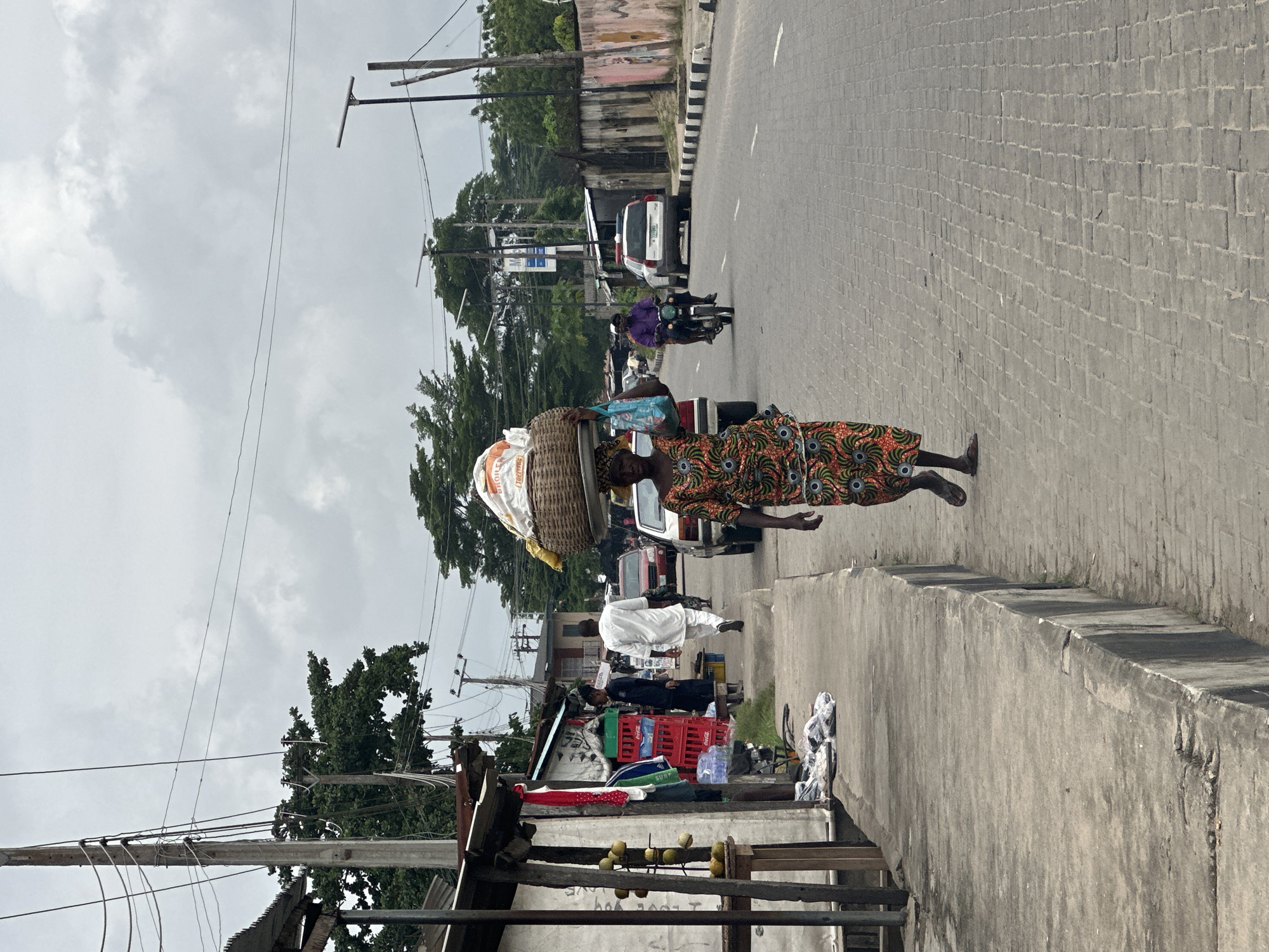 The image depicts a street scene in Badagry, Nigeria, under an overcast sky. In the foreground, a woman walks down a paved road, carrying a large woven basket filled with goods on her head. She is wearing a vibrantly patterned dress. To her left, a raised concrete sidewalk runs alongside a row of open-fronted market stalls. These stalls display various items including crates of drinks, fabric, and produce such as yellow fruits. A man in a white garment walks away from the stalls on the sidewalk. Further down the road, other people are visible, including a person on a motorcycle and several cars. The road is lined with trees and electrical poles and wires. The overall impression is of a busy, everyday market environment within a Nigerian town.