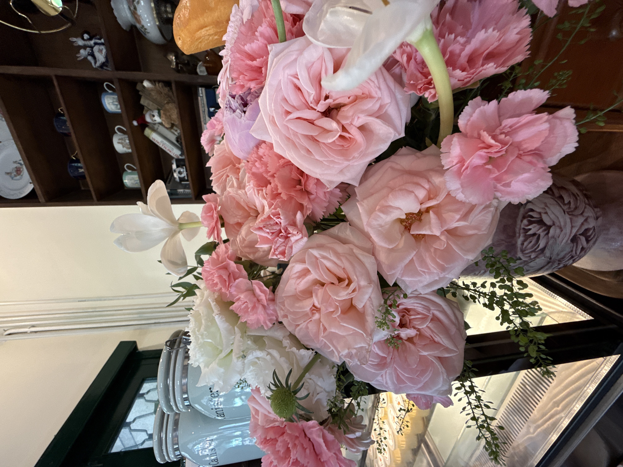 A close-up, high-angle shot of a bouquet with pink roses, white tulips, and carnations, set against a backdrop of a dark wooden shelf holding porcelain items. The bouquet is positioned in a glass vase atop stacked "Canister" labeled containers. Additional elements in the frame include a glass-encased counter or display case. The location is most likely Da Lat, Vietnam.