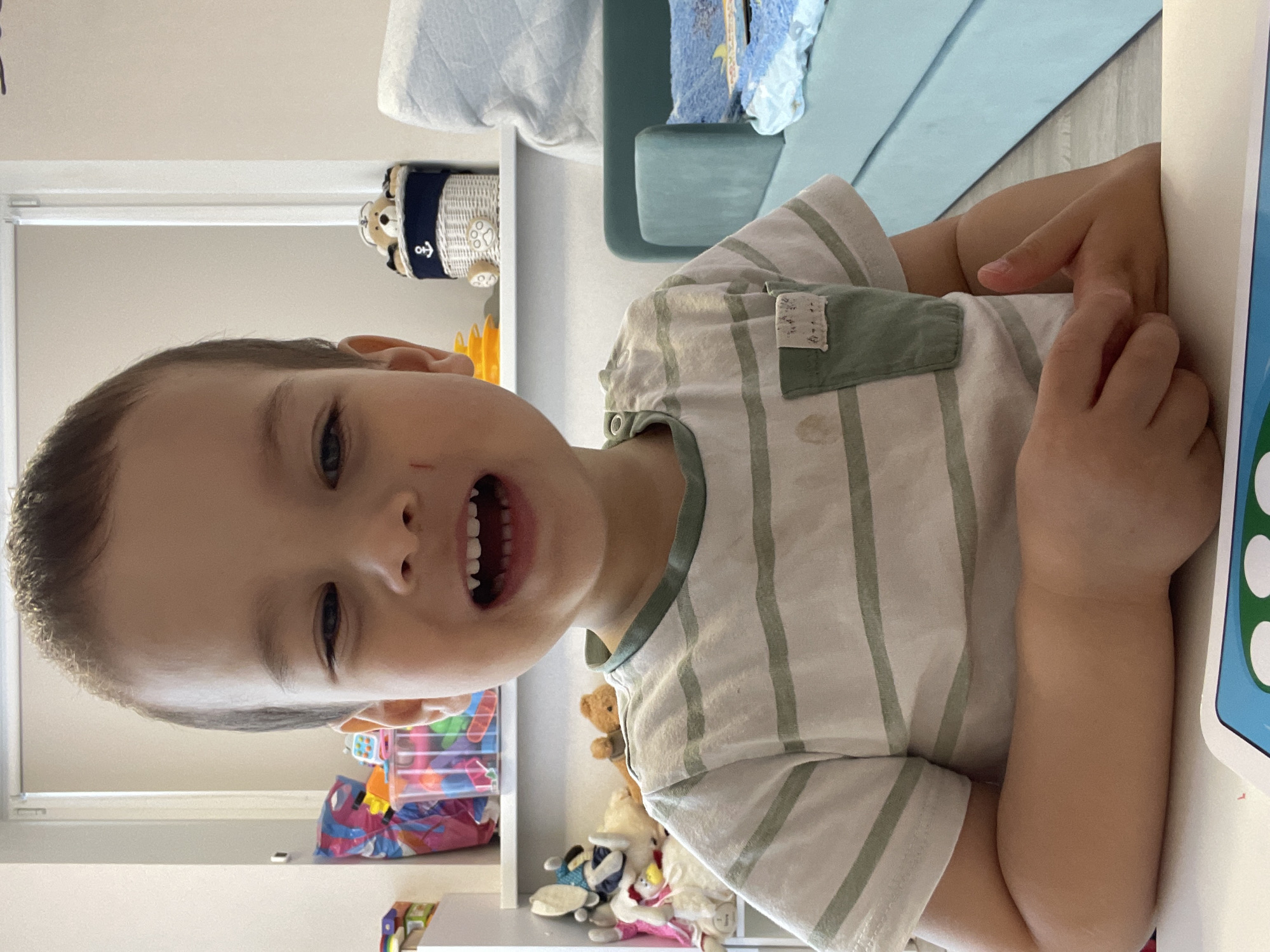 A young boy with short brown hair is smiling broadly, revealing his teeth. He is wearing a striped shirt with a small pocket. He is sitting at a table, possibly engaged with a tablet or similar device.
Behind him is a shelf unit with various items: toys, containers of objects, and decorative pieces. The background is blurred, suggesting a room in a home.
There is no definitive evidence indicating that this image was taken in Izhevsk, Russia.
