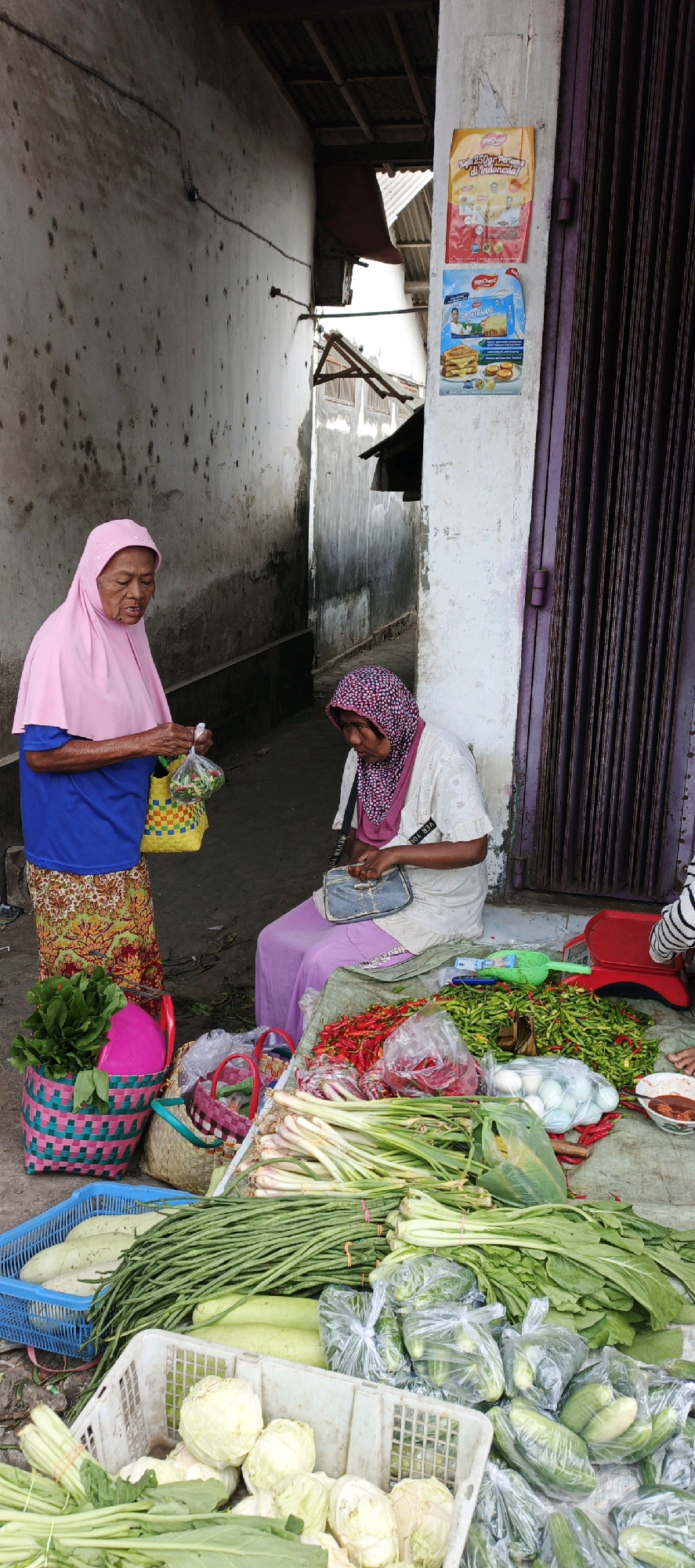 In the city of Kasiyan, Indonesia, two women are engaged in a transaction at a street market stall. The woman on the left, wearing a pink hijab and a blue shirt, is handing a plastic bag filled with items to the woman on the right. The woman on the right, also wearing a hijab and a white patterned top with a purple skirt, is seated and appears to be counting money or making a purchase. The stall is laden with a vibrant assortment of fresh produce, including red and green chilies, green beans, leafy greens, onions, cucumbers, and white cabbages. Baskets of various sizes and colors are used to display some of the vegetables. A red weighing scale and other market tools are visible. The background suggests an outdoor market setting with a textured wall and a corrugated metal roof overhead. Advertisements are visible on the wall behind the women.