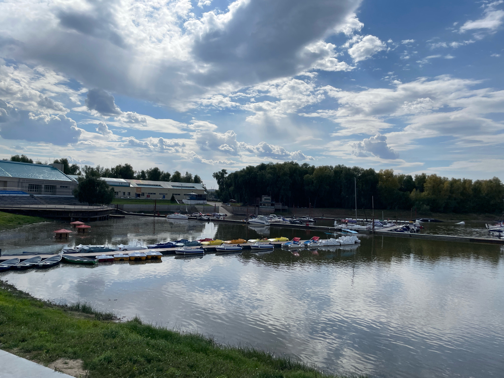 This image captures a view of the Irtysh River in Omsk, Russia, on a cloudy day. The foreground shows a body of water with reflections of the sky and boats. Along the embankment, a variety of small boats, including paddle boats and motorboats, are docked. In the middle ground, there are several buildings, some with awnings, and a tree line borders the riverbank. In the background, the sky is filled with dramatic clouds. There are no people visible in the image.