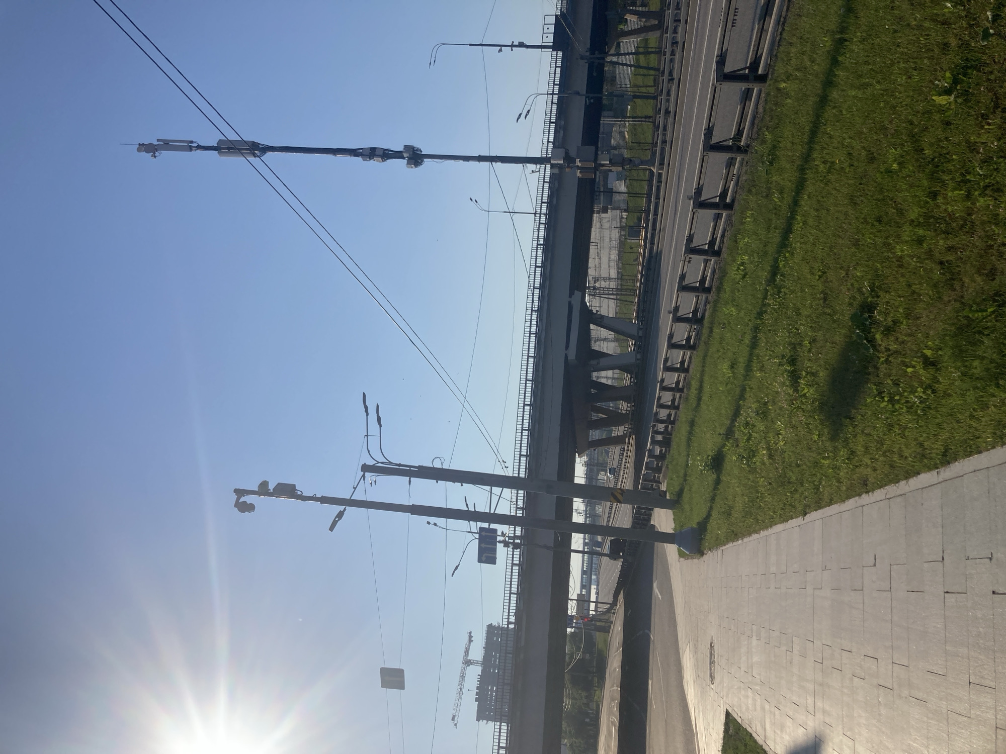 This is a full-shot, eye-level, outdoor photograph taken in Moscow, Russia, under a clear blue sky with a bright sun causing lens flare on the left side of the image. The scene depicts a modern cityscape featuring an elevated highway, a pedestrian walkway, and infrastructure elements like streetlights and communication towers.
In the foreground, a paved pedestrian walkway made of light-colored rectangular tiles runs along the right side of the image, bordered by a strip of well-maintained green grass. The grass extends to the edge of the roadway.
The elevated highway is a prominent feature, crossing the scene horizontally. It has a concrete barrier along its sides and appears to have multiple lanes of traffic, although only a few vehicles are visible, including a black car in the distance. The highway's structure includes concrete pillars supporting the elevated road.
Two tall communication towers are visible, one on the right and another, shorter one on the left. The tower on the right is significantly taller and equipped with multiple antennas and equipment. The tower on the left also has antennas and what appears to be a surveillance camera.
In the background, to the left, a construction site is visible with a large crane and a partially built structure. Several overhead power lines stretch across the sky, connecting to the communication towers. A few streetlights are also positioned along the walkway and near the highway. The overall impression is one of urban development and infrastructure in a well-lit environment.