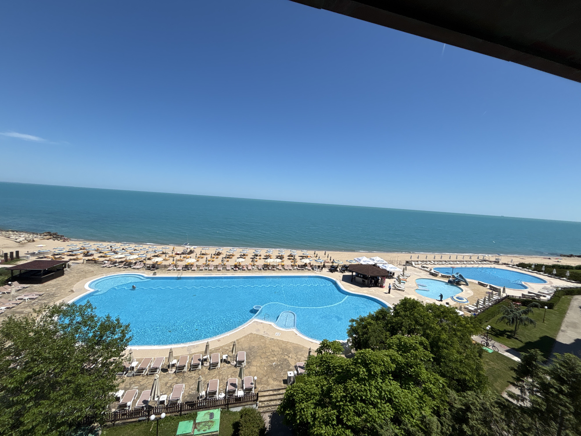 The image shows a resort area in Varna, Bulgaria, featuring multiple swimming pools and a beach. The main pool is a large, irregularly shaped body of bright blue water with a person swimming in it. Adjacent to this pool is a smaller, shallow pool with a slide. The pools are surrounded by a paved patio area with numerous lounge chairs and umbrellas.
Beyond the pools, a sandy beach is visible, lined with rows of lounge chairs and umbrellas. The sea, a vibrant turquoise color, stretches out to the horizon under a clear blue sky. To the left of the main pool, there is a thatched-roof structure that appears to be a bar or cabana. Green trees and manicured lawns are present in the foreground and to the right, indicating a well-maintained resort environment. In the distance, there is a hint of a rocky shoreline. The overall impression is one of a sunny, recreational destination.
