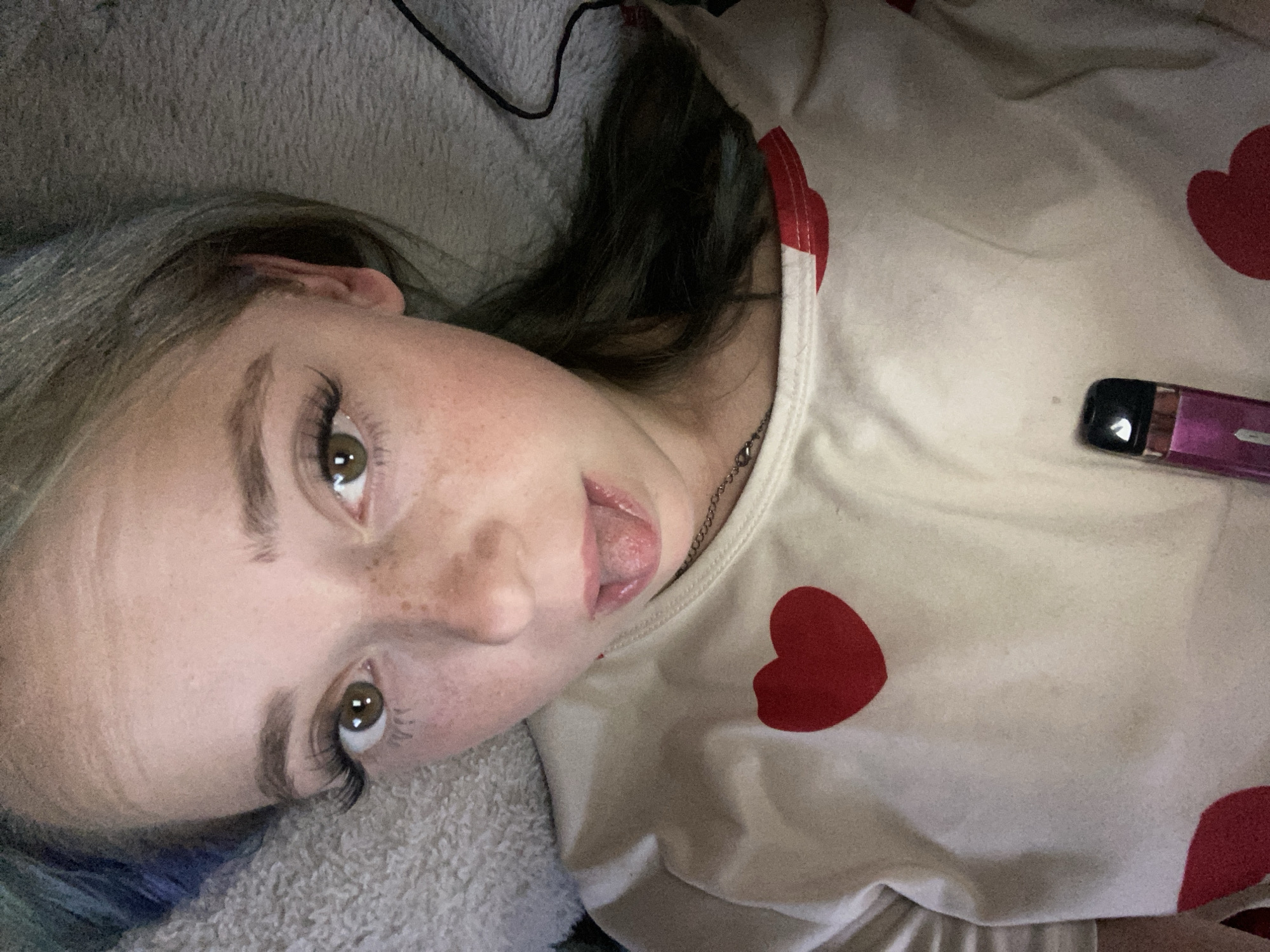 The media file is a close-up, vertically oriented, indoor photograph of a young person lying down. The subject has light skin with freckles, brown eyes, and dark hair that transitions to a blue hue. They are wearing a cream-colored shirt adorned with red hearts. Their tongue is slightly extended, and they are looking towards the camera with a neutral expression. To the right of the subject's chest, a small, pink electronic device with a black tip is visible on the shirt. The background consists of a soft, textured gray blanket. The lighting appears to be artificial, suggesting an indoor setting, possibly a bedroom. There is no visible text in the image.