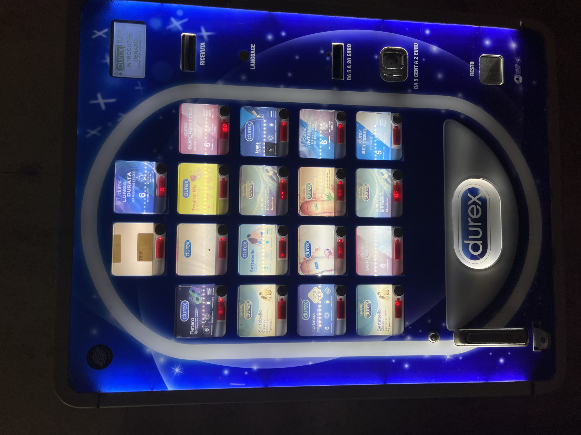 The image depicts a close-up of a condom vending machine. The machine is illuminated and features a variety of Durex condom products. Text on the machine is in Italian, including instructions to insert money and language options. The machine accepts coins ranging from 5 cents to 2 euros, and banknotes from 5 to 20 euros. The selection of condoms includes various types, such as "Lunga Durata," "Tropical Mix," "Jeans," "Massage 2in1," and "Settebello." Each selection shows the price.
The machine is likely located in Aristanis/Oristano, Italy, given the Italian language on the machine and the general prevalence of such vending machines in European countries. However, without further contextual information, it is impossible to determine the precise location.