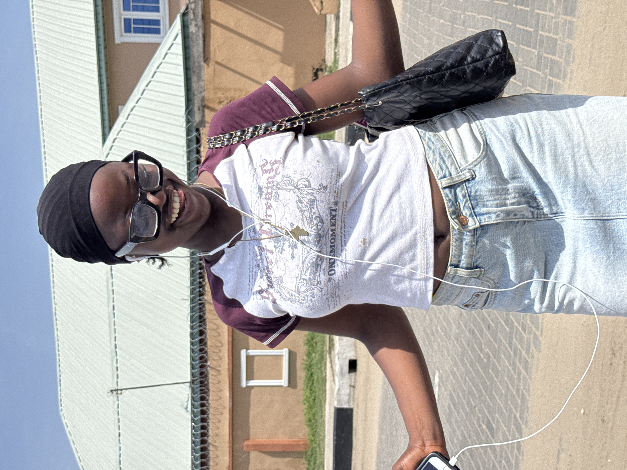 A woman stands on a paved surface near a building with corrugated metal siding in Badagry, Nigeria. She is wearing a black head covering, sunglasses, a white t-shirt with burgundy sleeves, and light blue jeans. She has a black quilted purse slung over her shoulder. Earphone cords are visible, connected to a device in her hand. She is smiling.