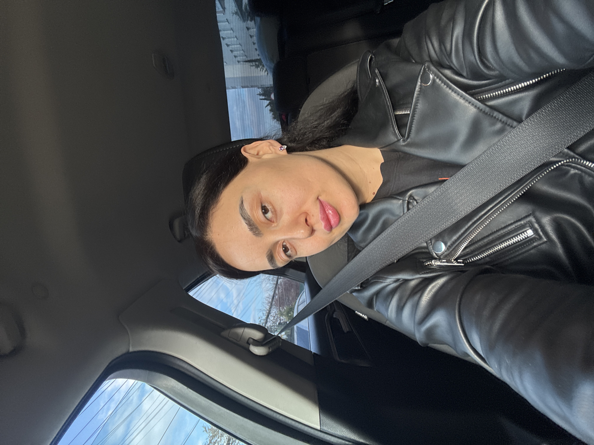 A woman is in the driver's seat of a car. She is wearing a black leather jacket and a seatbelt. The car appears to be in motion, with a glimpse of buildings and sky visible through the window. The location is unknown.